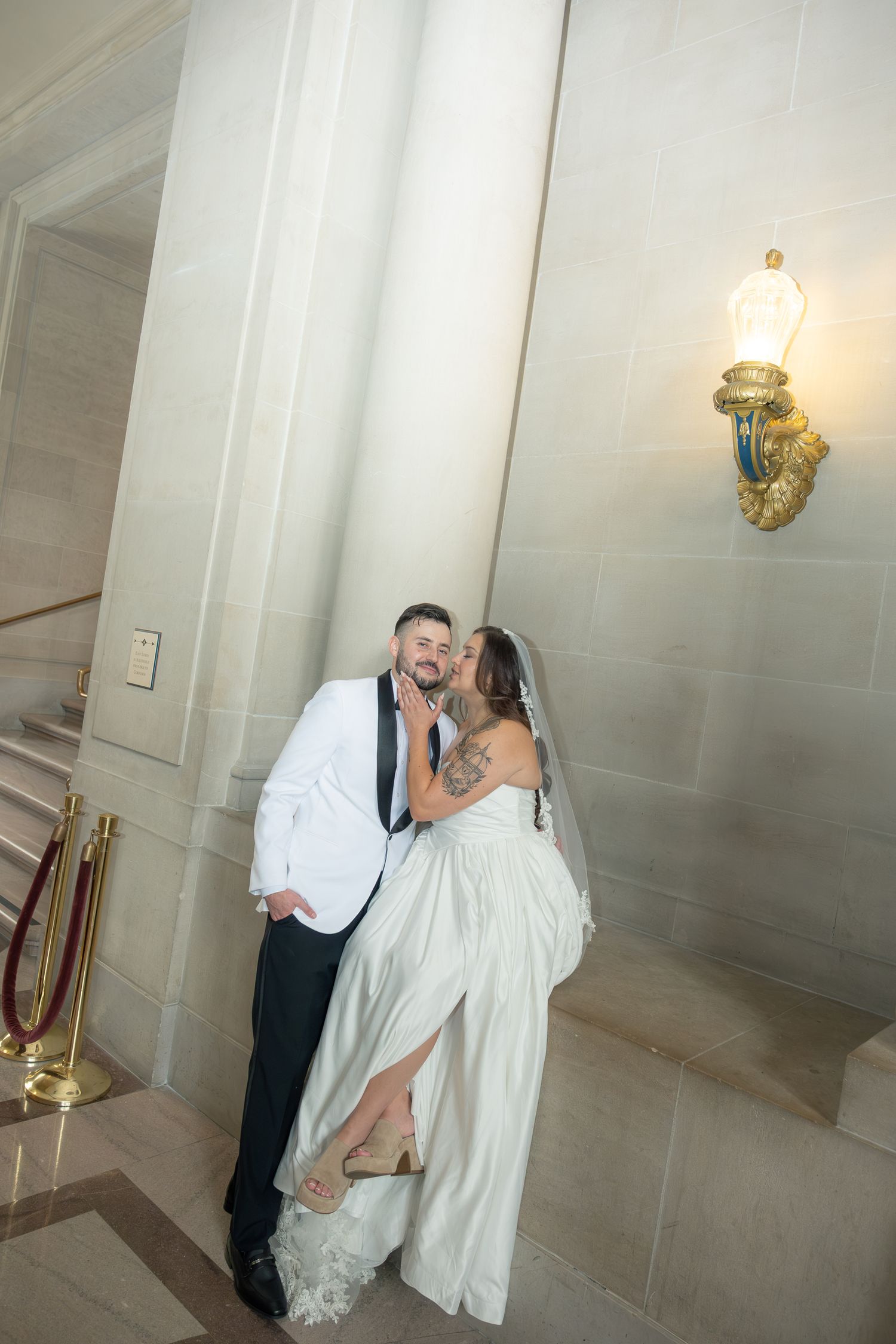 Newlyweds sharing a kiss near a marble column with golden wall sconce.