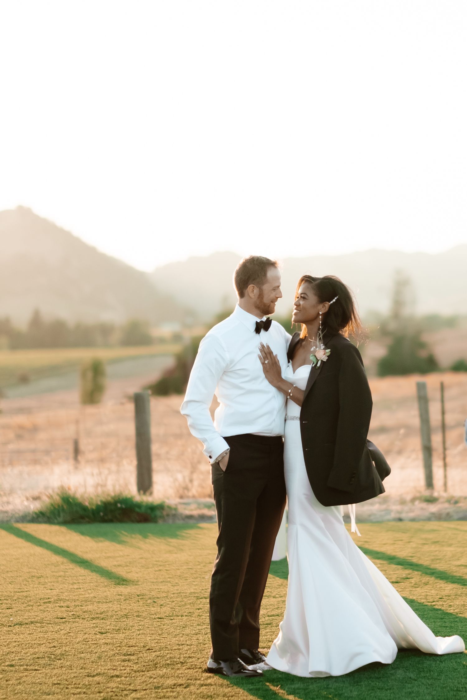 Interracial newlywed couple share romantic sunset moment on green lawn with mountain backdrop.