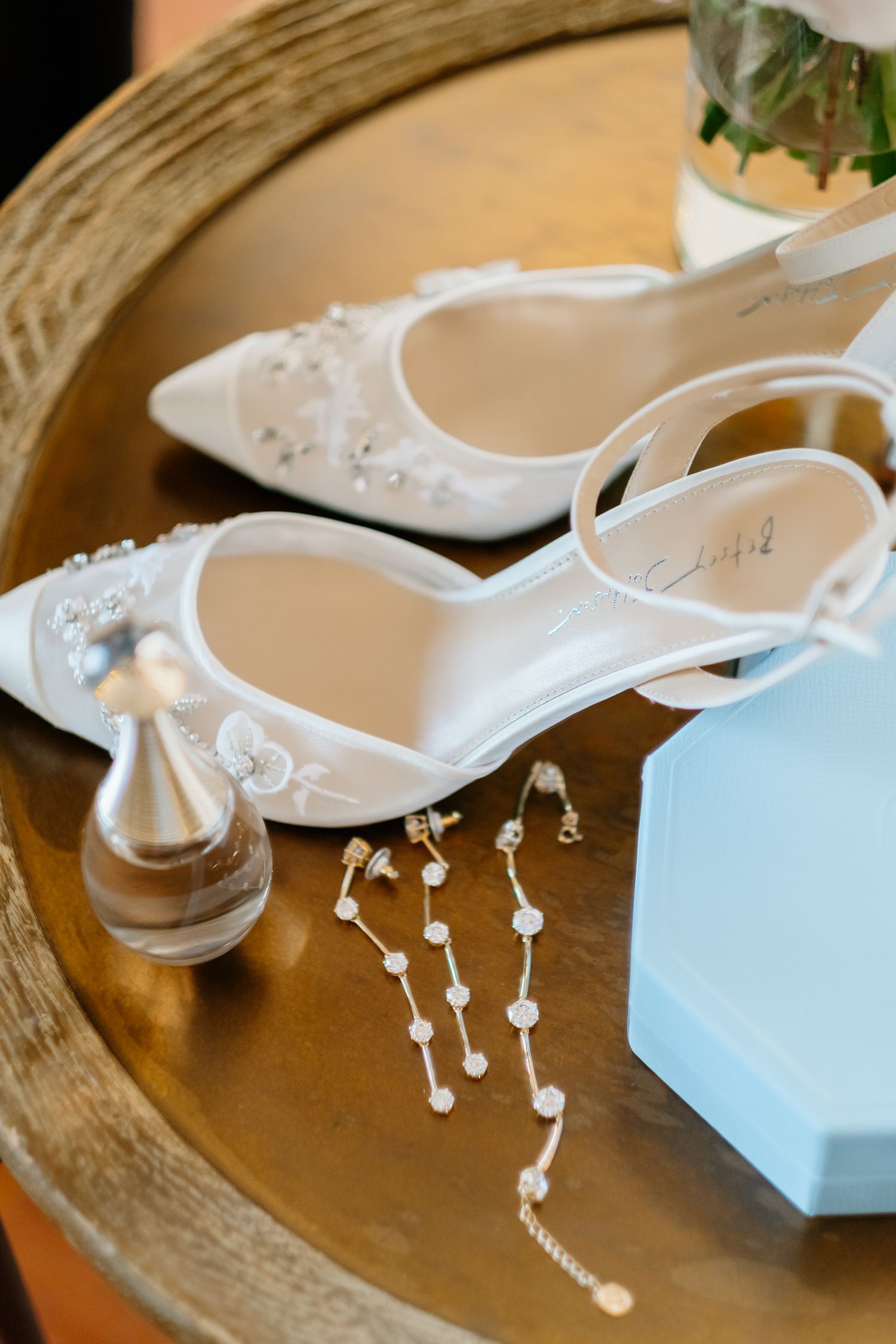 White bridal heels with pearl embellishments beside perfume bottle and pearl jewelry on wooden table.
