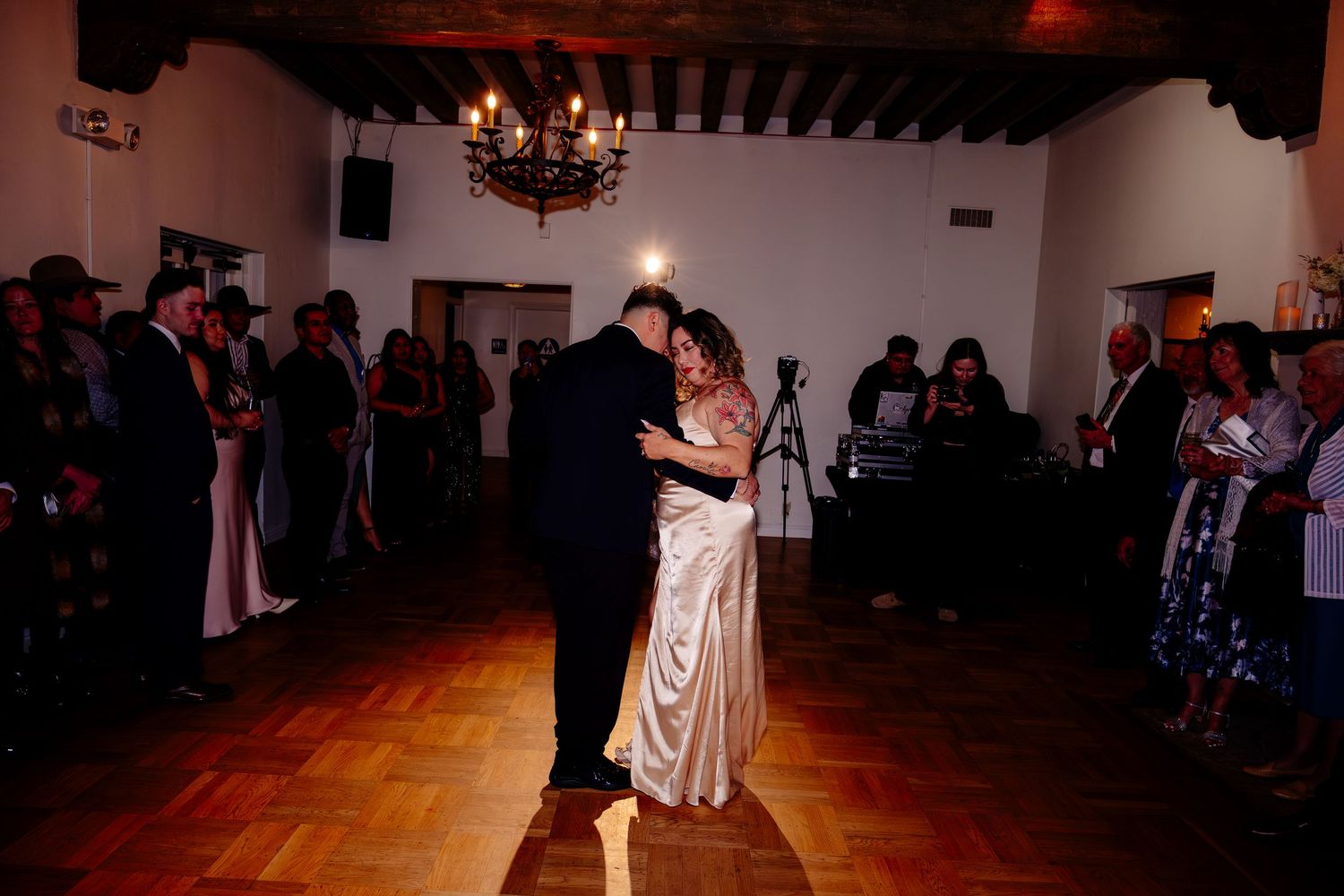 Couple shares first dance in rustic wedding venue with chandelier and guests watching.