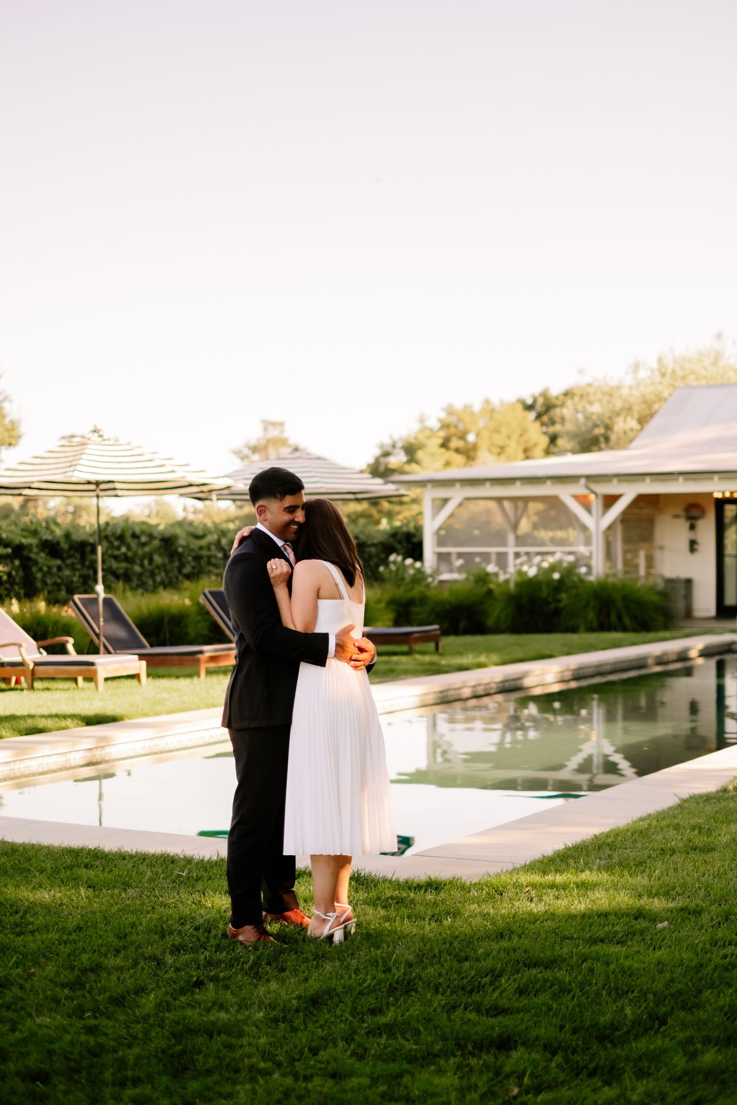 Bride and groom embracing romantically by a serene pool at golden hour in elegant attire.