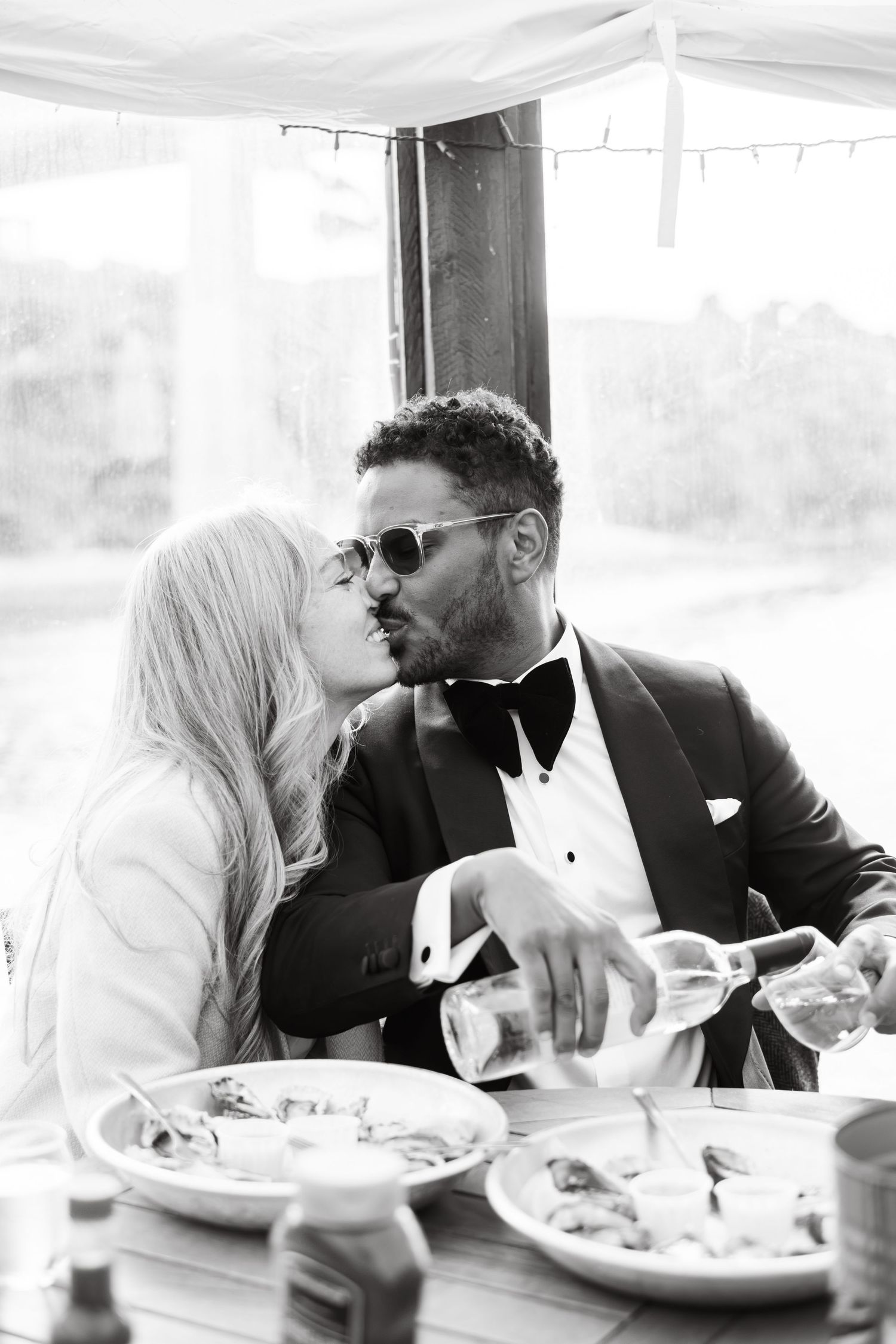 Black and white photo of a couple sharing a kiss at a wedding reception dinner table, groom in tuxedo.