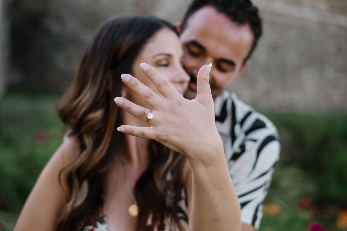 Couple celebrates engagement, woman shows off diamond ring while partner kisses her cheek outdoors.