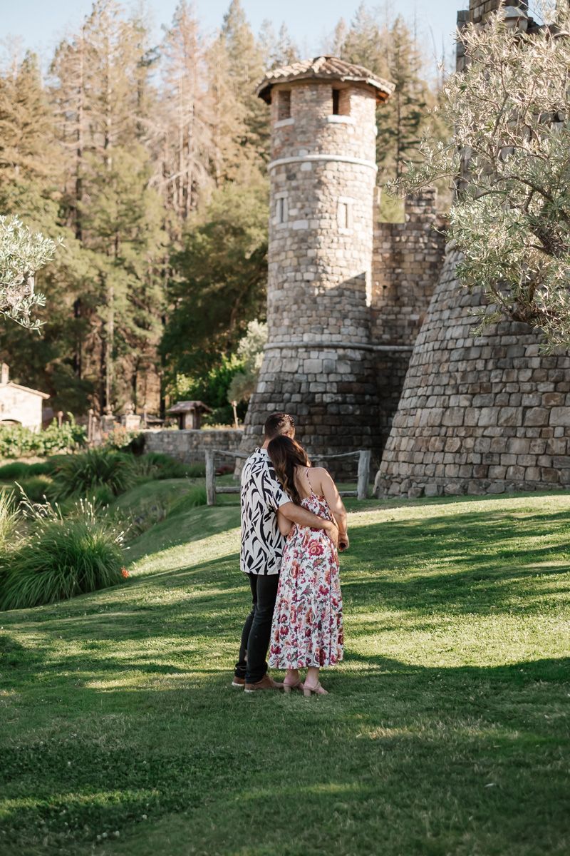 Engaged couple embracing on green lawn beside a rustic stone tower winery.