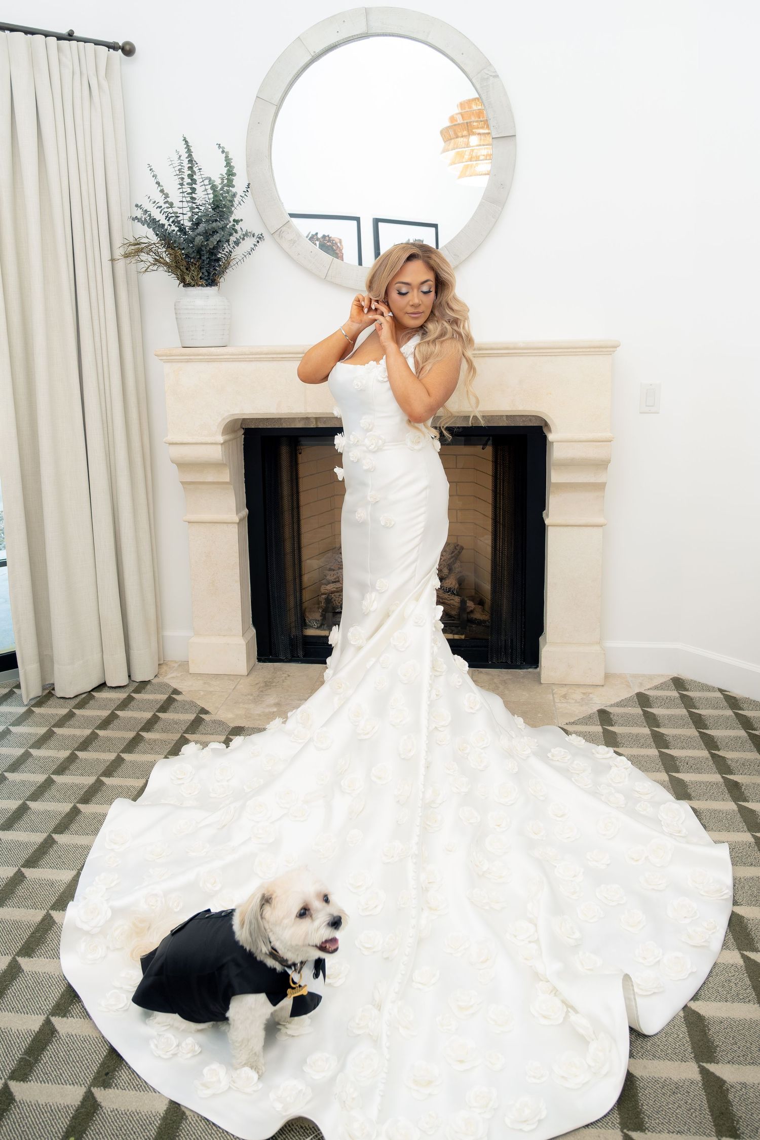 Bride in floral mermaid gown adjusting hair while small dog in tuxedo sits on patterned floor.