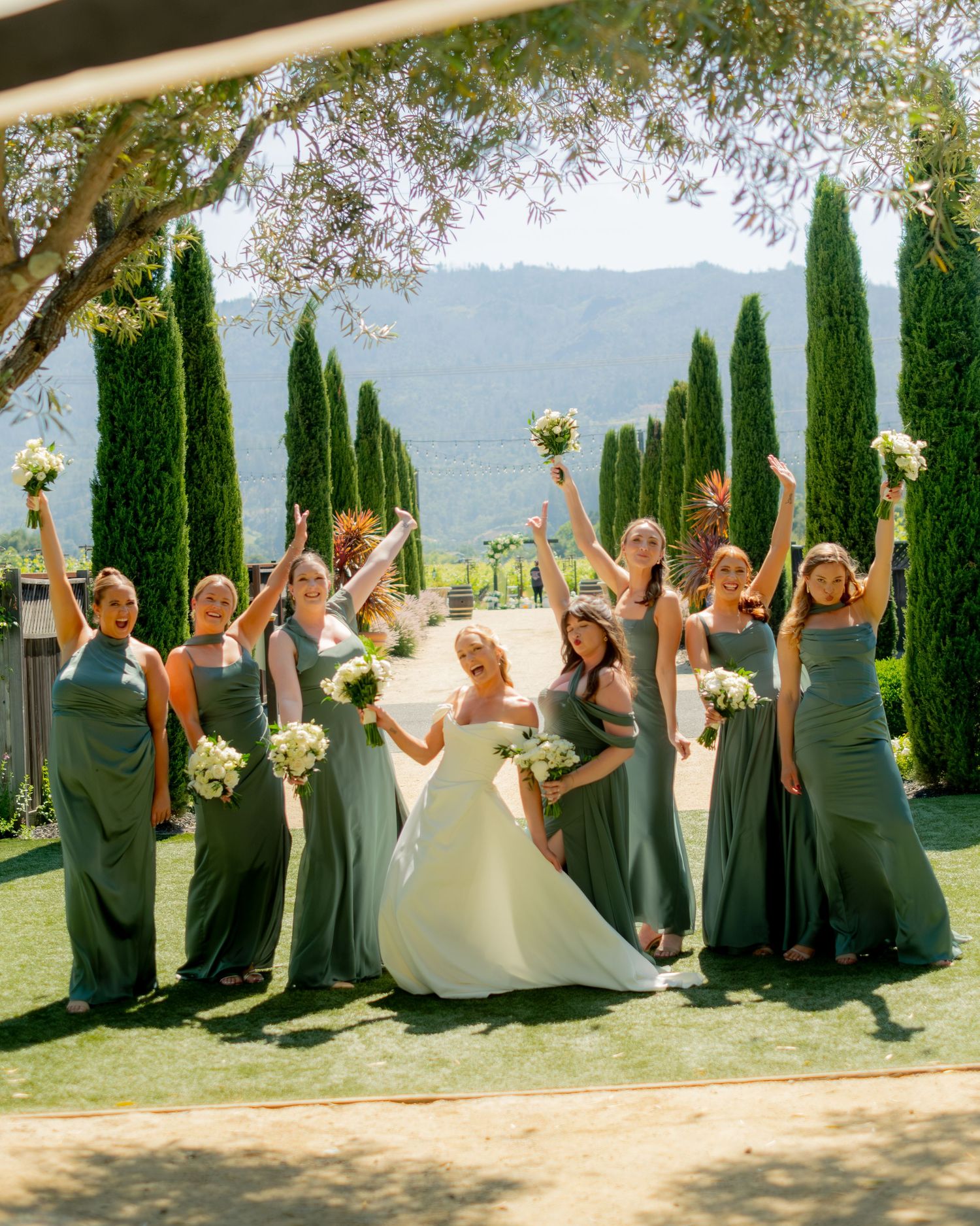 Bride and bridesmaids in sage green gowns celebrate joyfully with bouquets at scenic vineyard wedding.