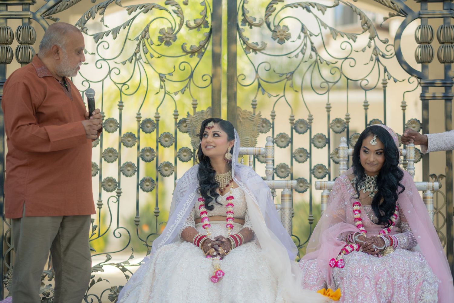Two Indian brides in white and pink lehengas seated before ornate golden gates.