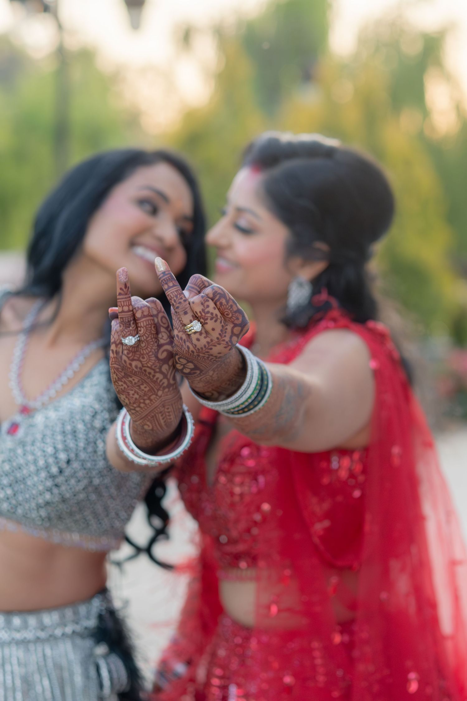 Two women in Indian wedding attire showing henna hands and engagement rings outdoors.