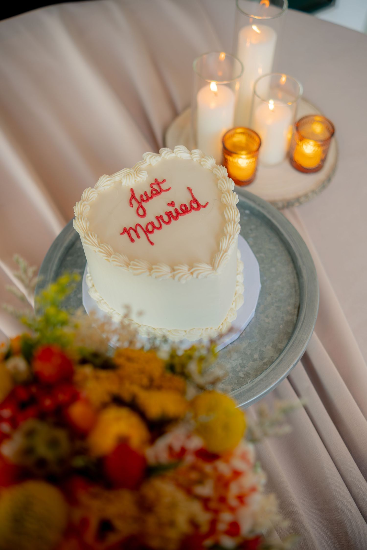 Heart-shaped white wedding cake inscribed with Just Married in red frosting beside candles and colorful flowers.
