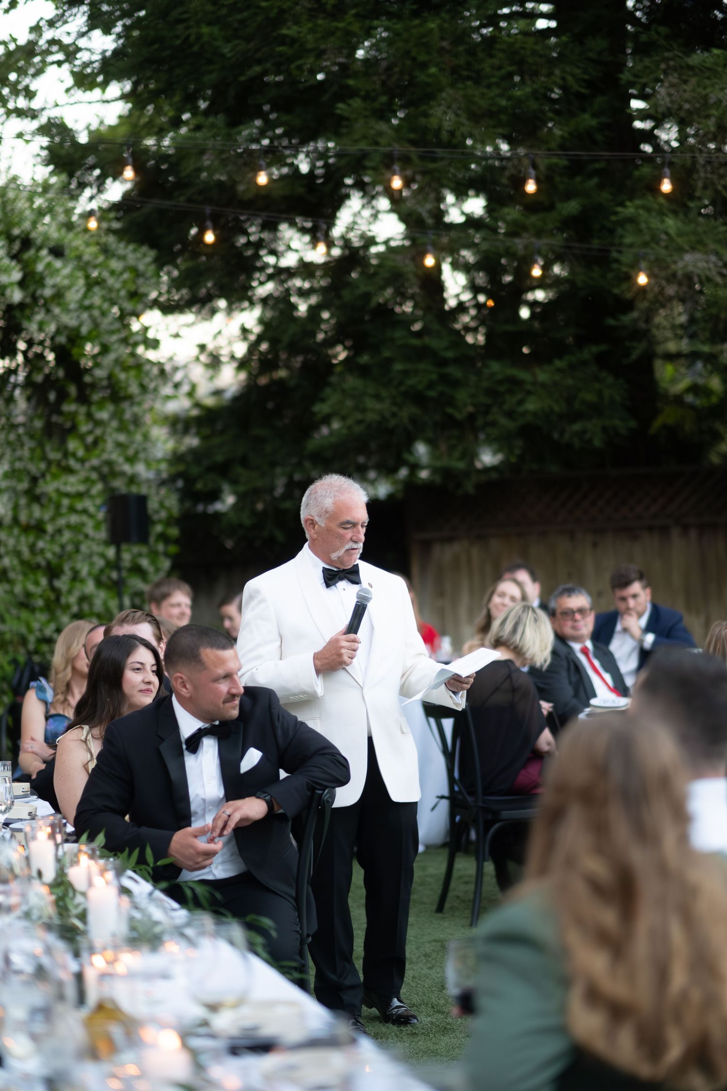Older gentleman in white tuxedo jacket giving a heartfelt speech at an outdoor wedding reception dinner.