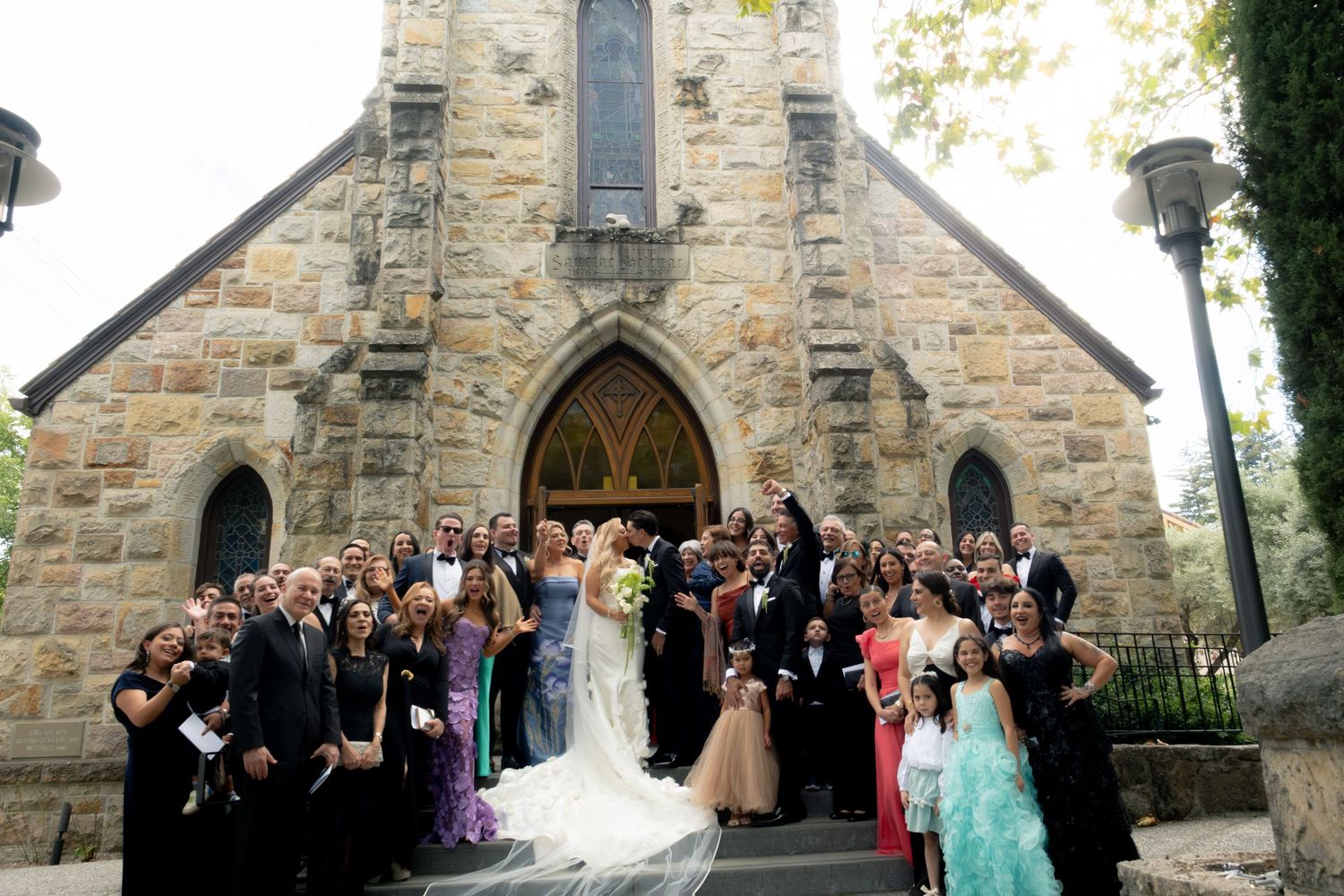 Large wedding party posing outside a historic stone church after ceremony.