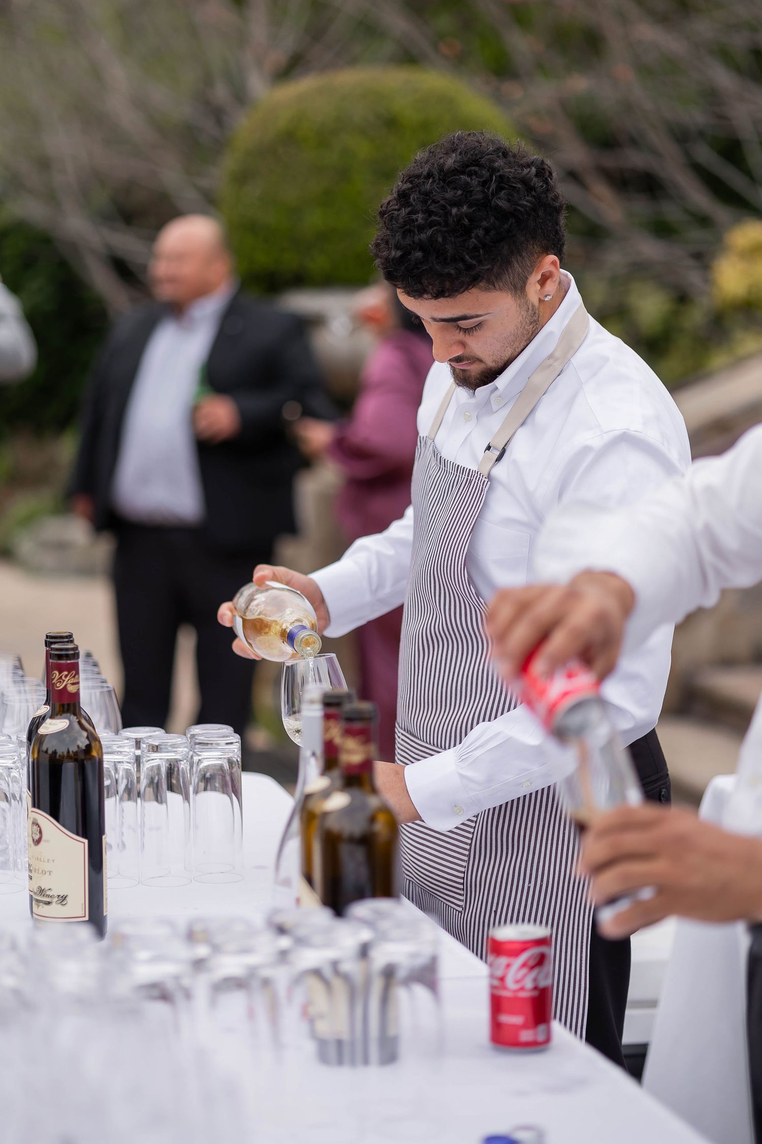 Bartender in striped apron pouring drinks at an outdoor wedding cocktail hour bar.