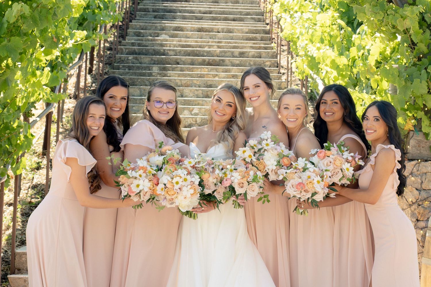 Bride with six bridesmaids in blush dresses holding colorful bouquets on vineyard stone staircase.