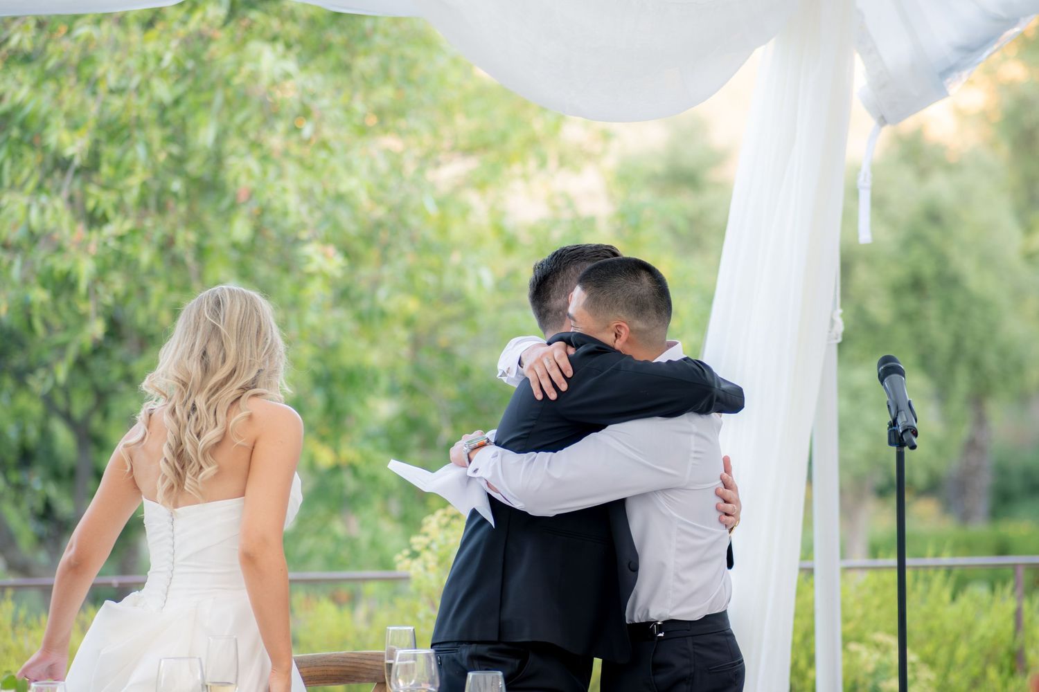 Groom sharing heartfelt hug with officiant during outdoor wedding ceremony as bride watches nearby.