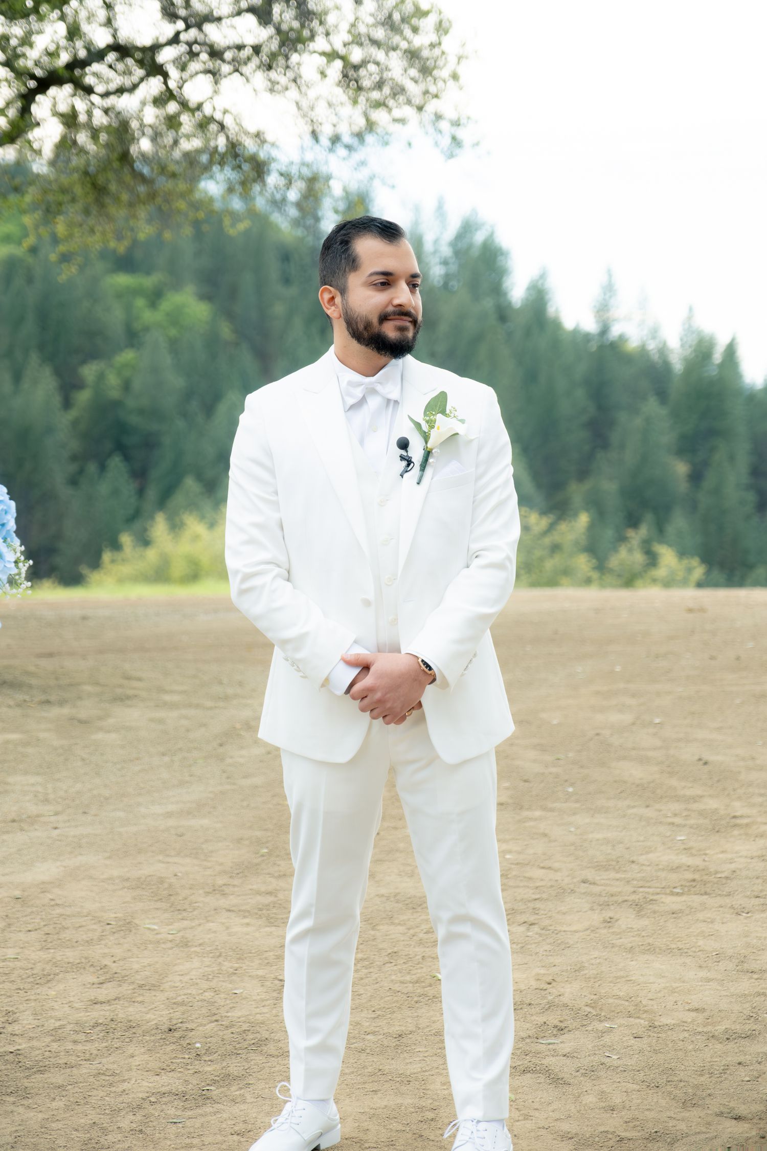 Groom stands at outdoor wedding altar in white suit with boutonniere, forest backdrop.