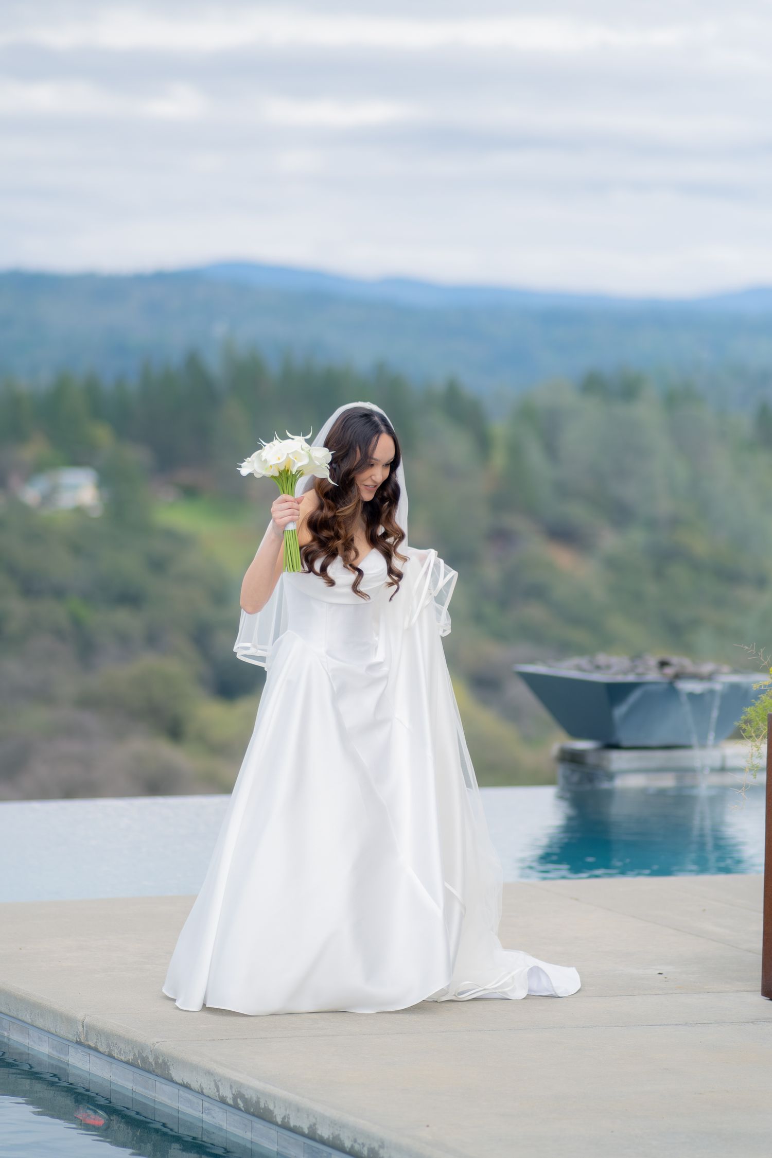 Bride in white gown holding bouquet by infinity pool with mountain views.