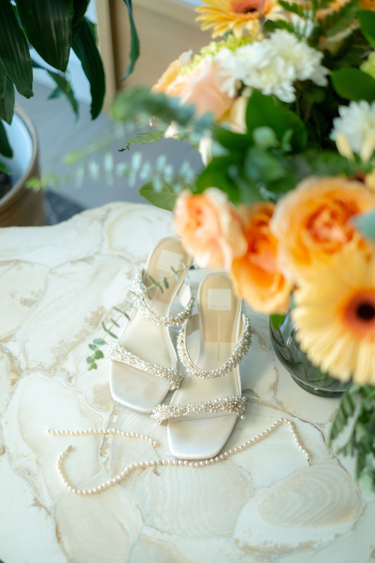 Elegant white bridal heels adorned with pearl straps displayed on marble surface near orange floral bouquet.