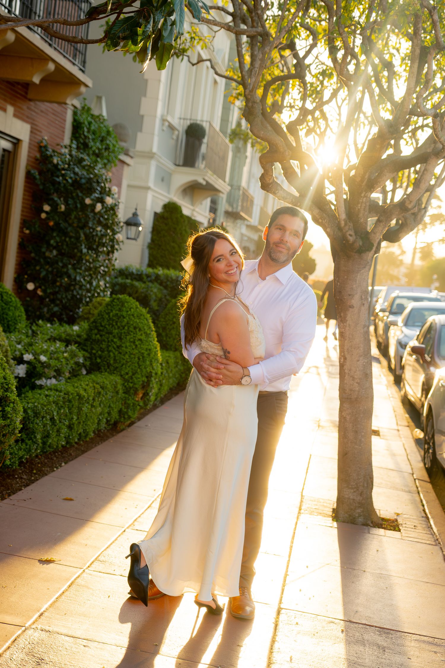 Couple posing together on tree-lined sidewalk at sunset, woman in cream dress smiling with man behind her.