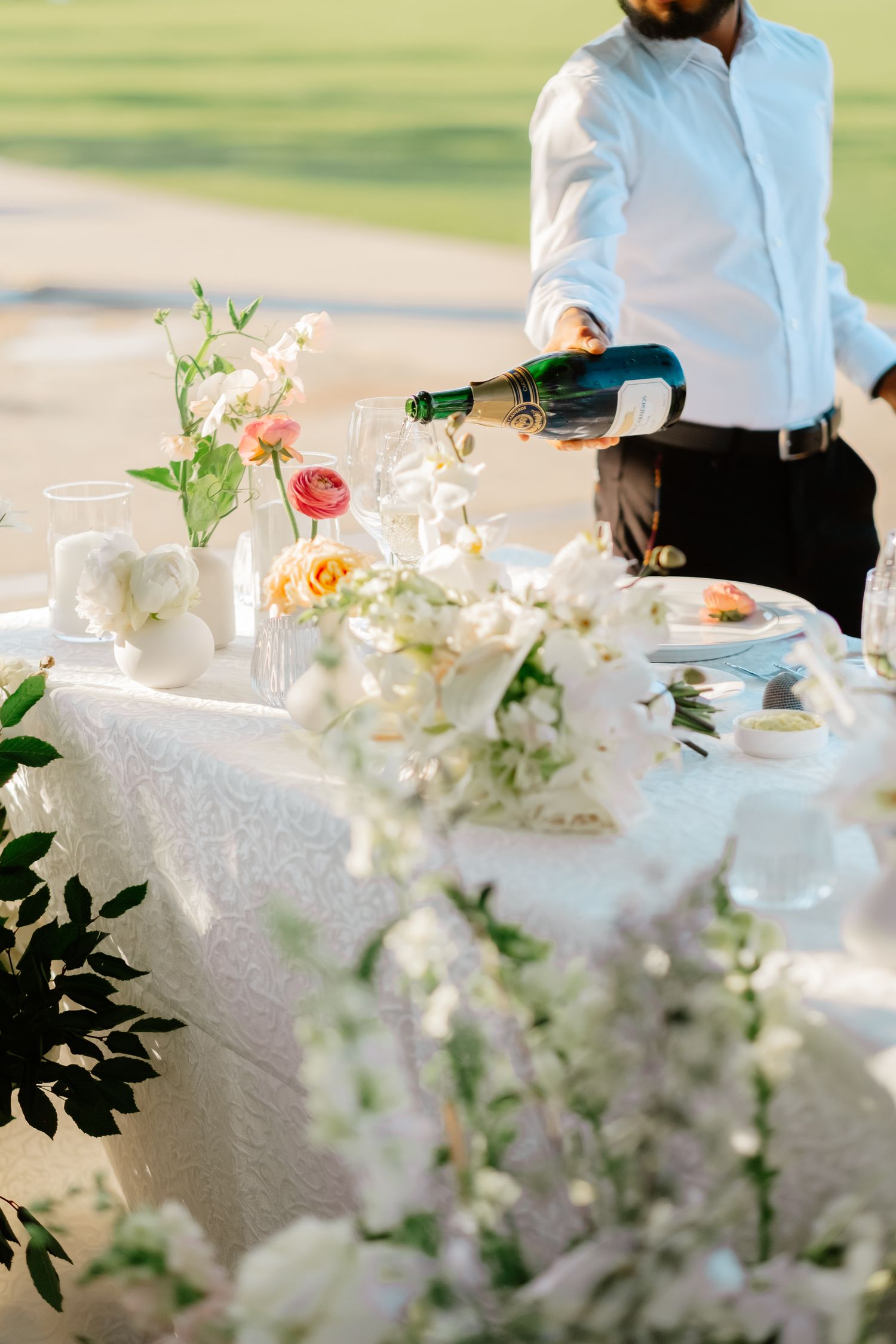 Waiter pouring champagne at a beautifully decorated wedding sweetheart table with white floral arrangements and candles.