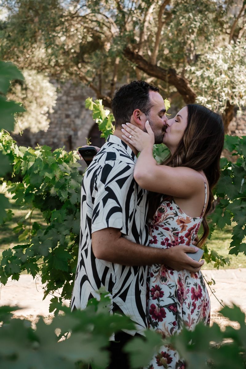 Couple sharing a passionate kiss surrounded by green grapevine leaves in a vineyard.