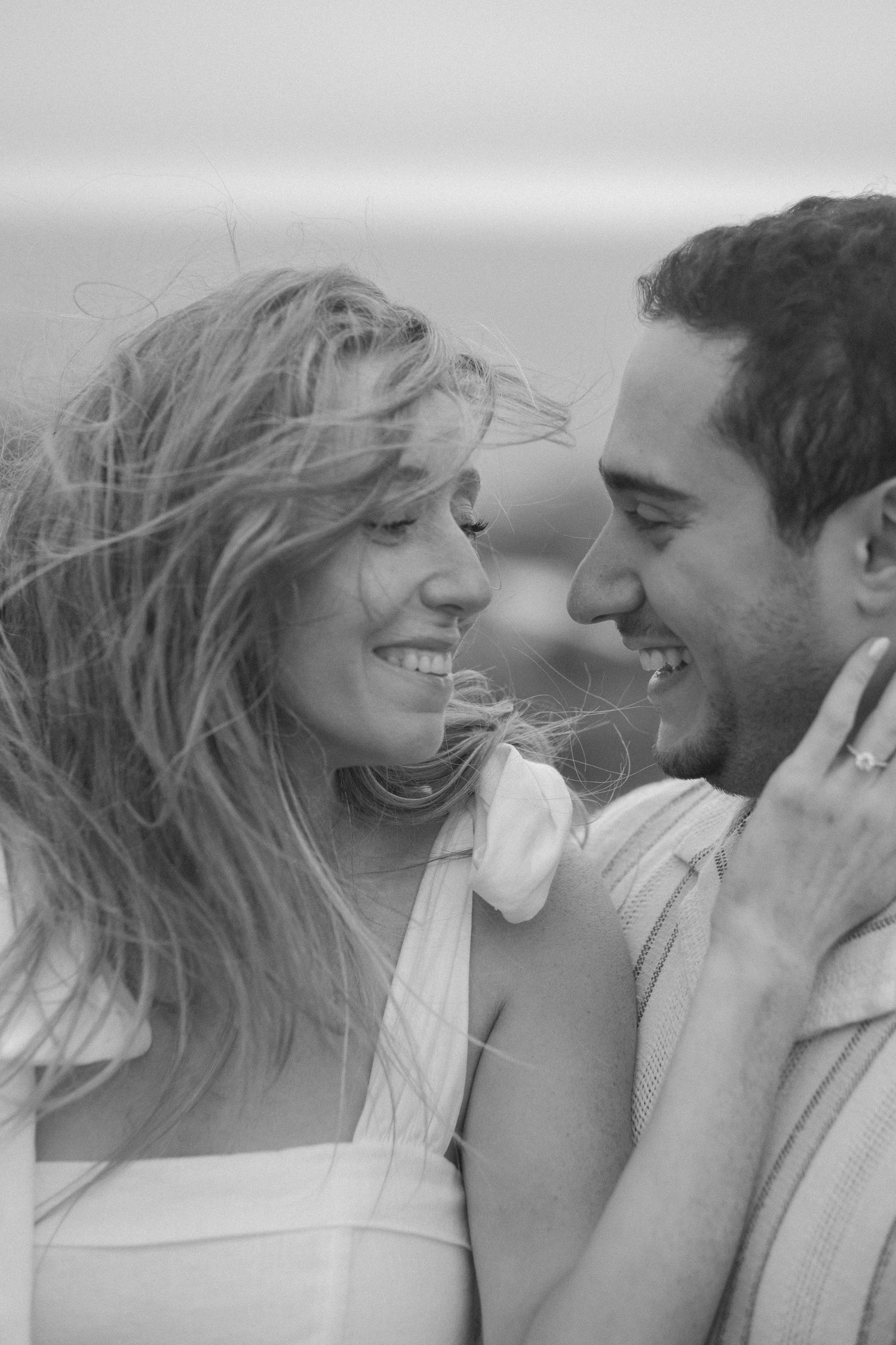 Black and white close-up of a smiling couple facing each other with windswept hair at the beach.