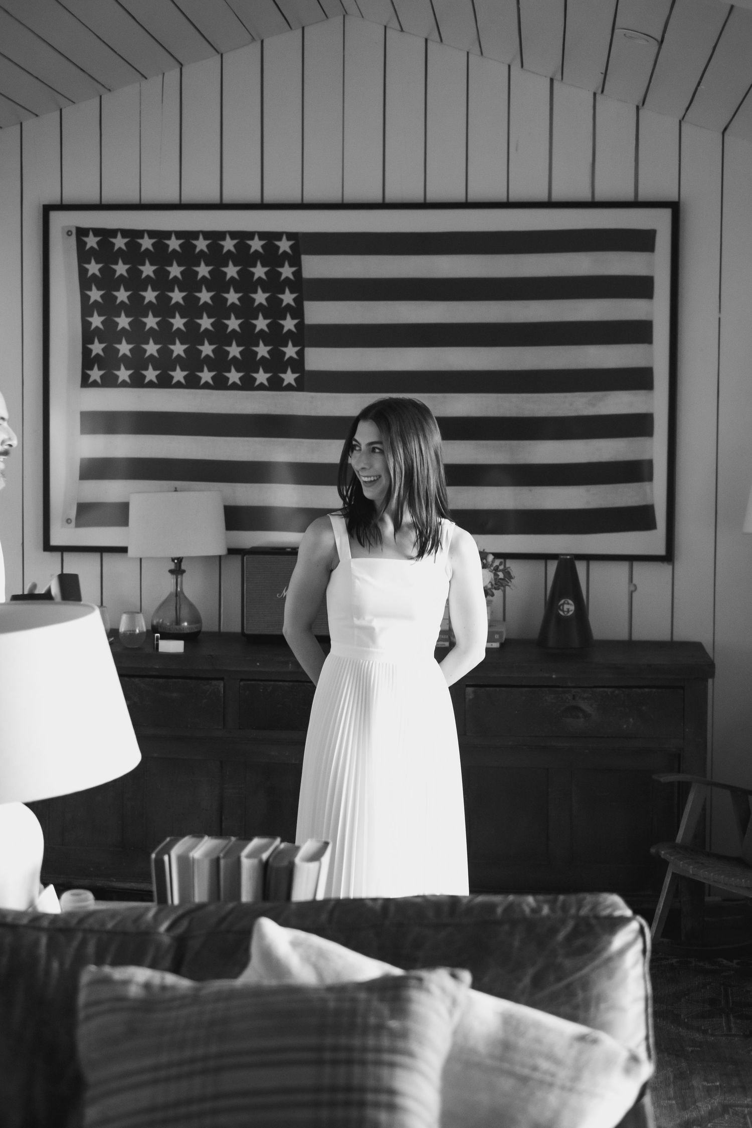 Black and white photo of bride in white pleated dress standing before large American flag wall art.