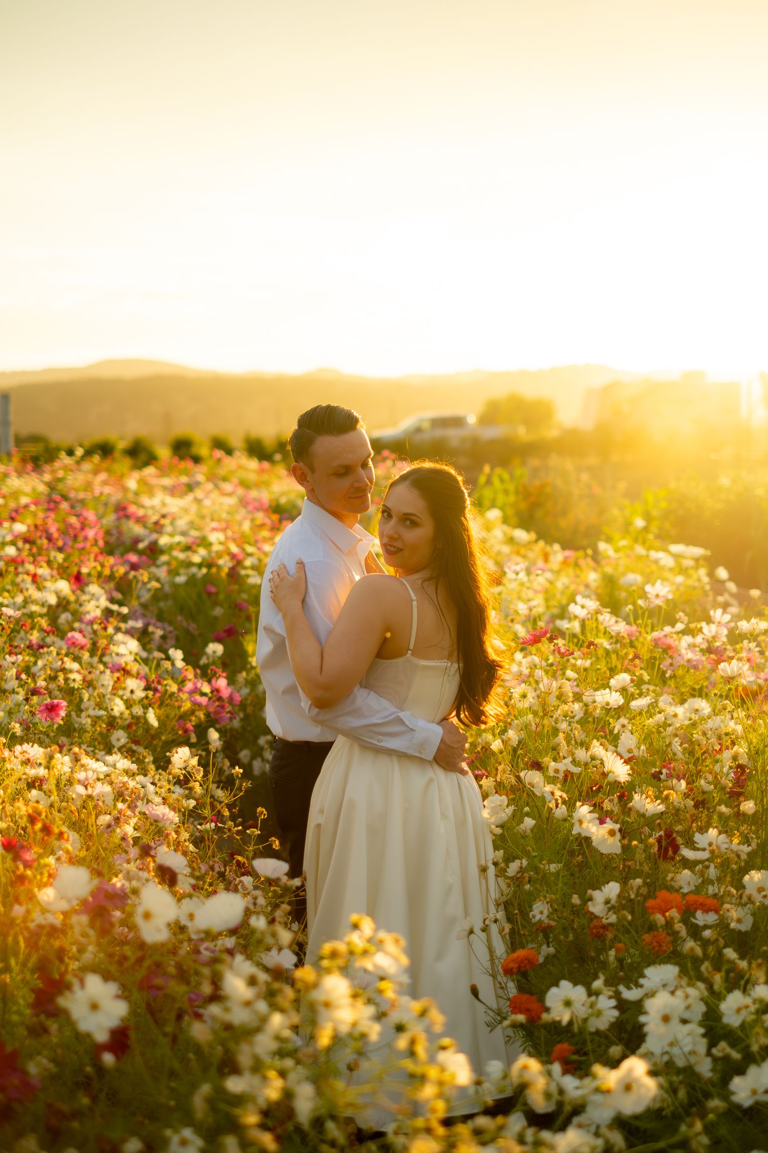 Couple embracing in a vibrant flower field at golden sunset, woman in white dress, romantic outdoor portrait.