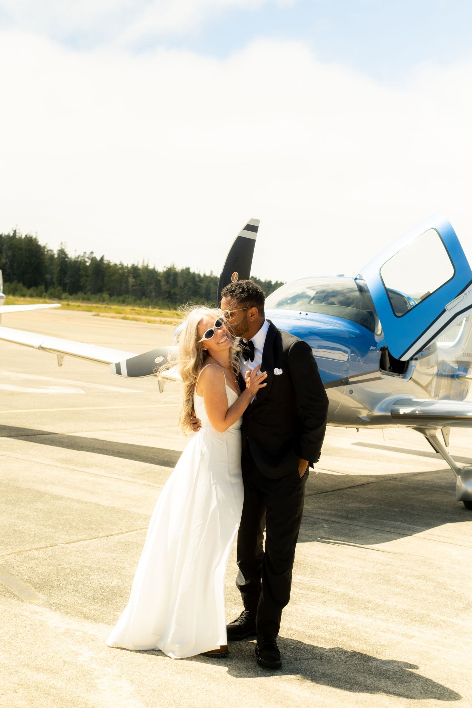 Bride and groom kissing in front of a blue propeller plane on a sunny airport tarmac on their wedding day.