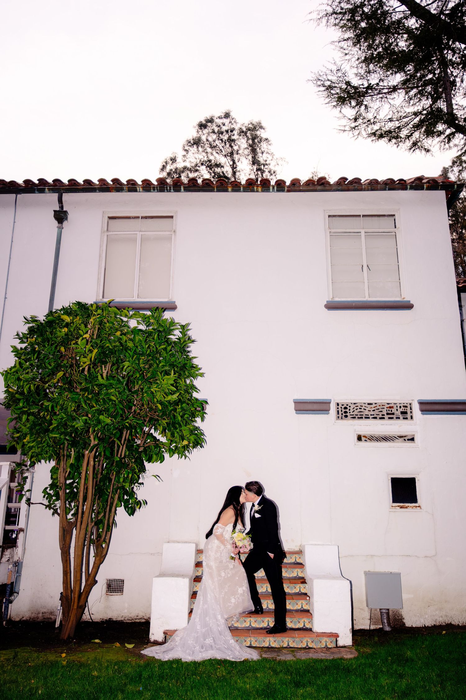 Bride and groom share a kiss on steps beside a white building during wedding portraits.
