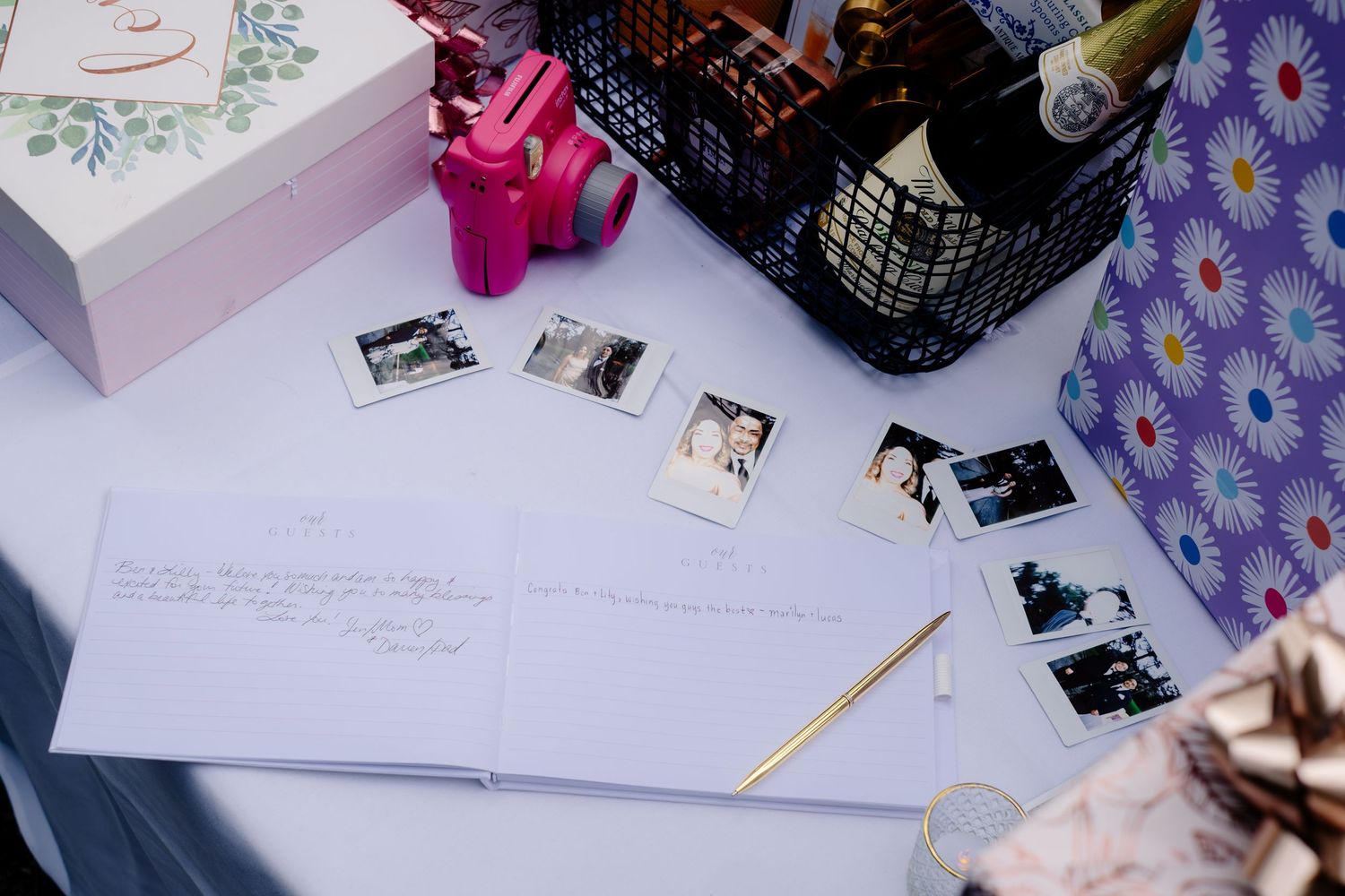 Wedding guest book table with Polaroid photos, gold pen, gift boxes, champagne bottle, and floral wrapping paper.