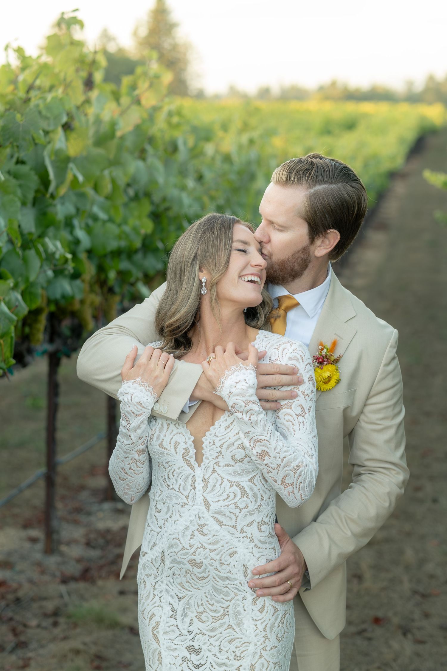 Groom embraces bride from behind with a kiss in a lush vineyard at golden hour.