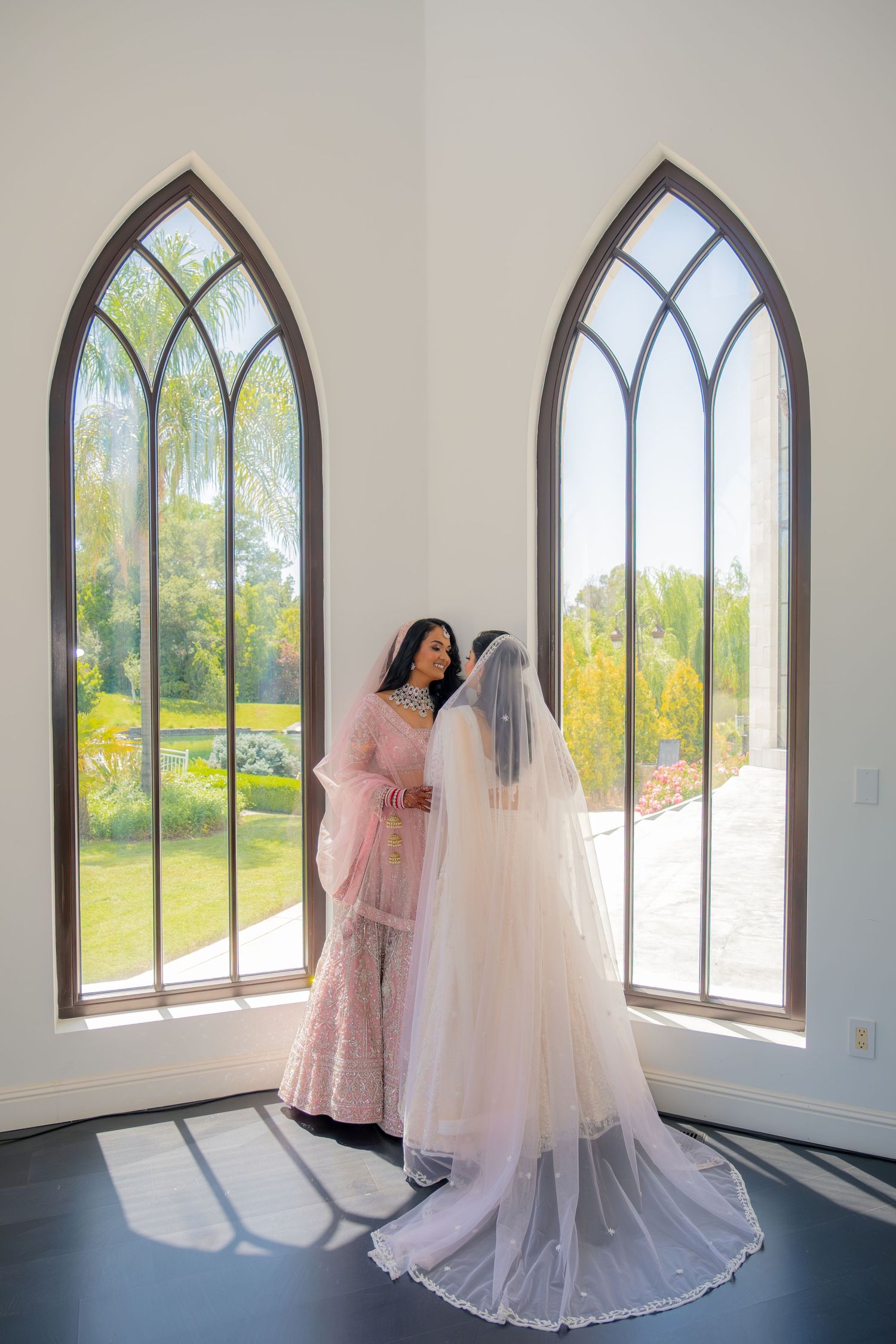 Bride in white gown with long veil sharing intimate moment with woman in pink near chapel windows.