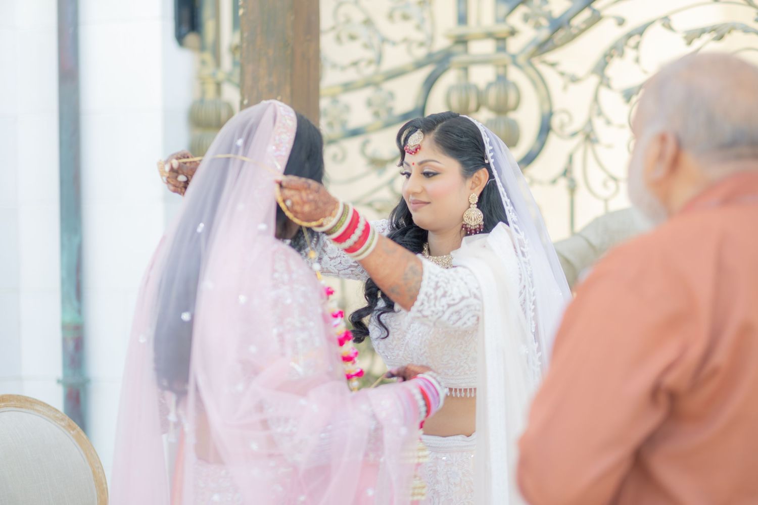 Indian bride in white lehenga receives help adjusting her veil during traditional wedding ceremony preparations.