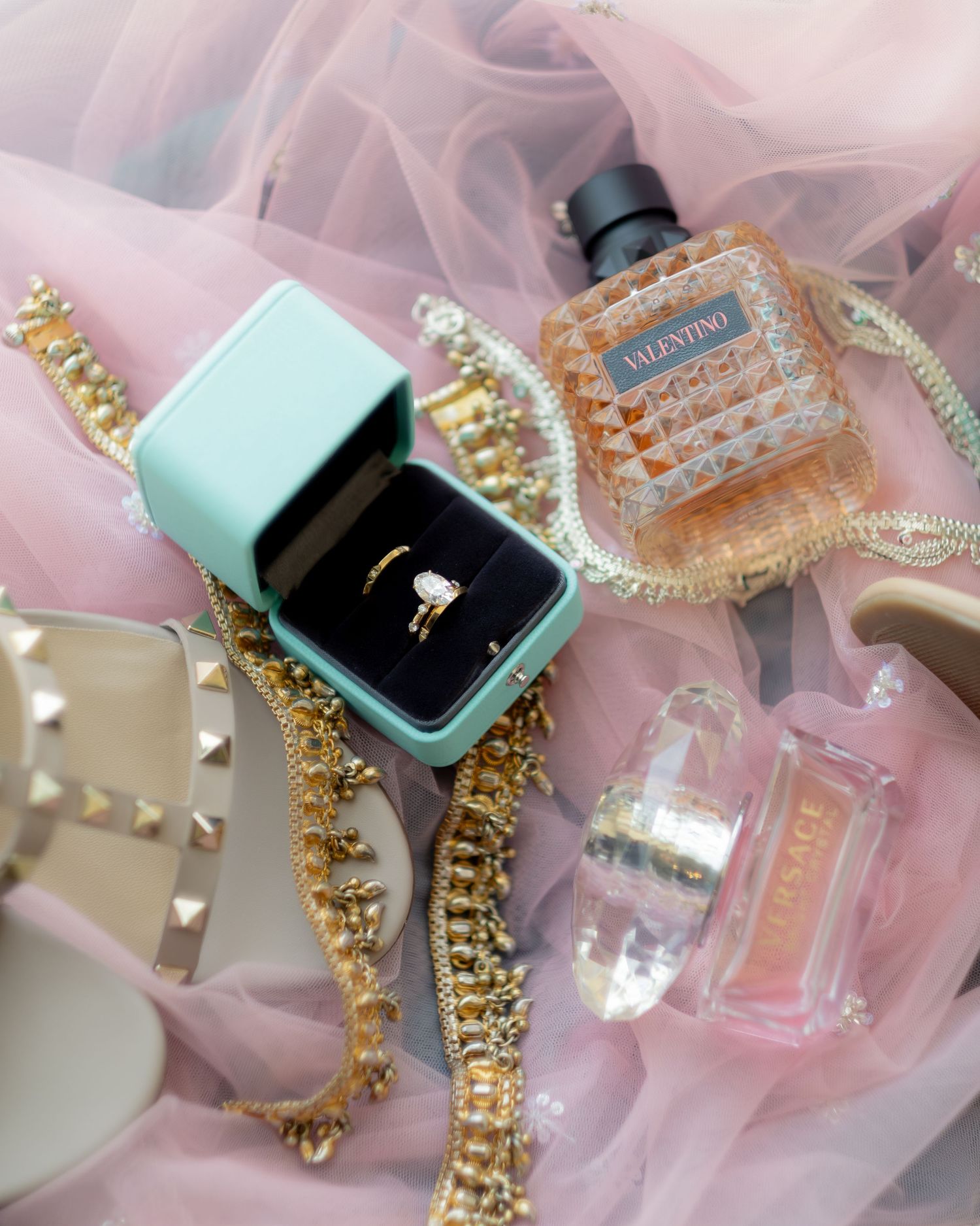 Flat lay of diamond ring in mint box, Valentino perfume, gold jewelry, and studded heels on pink tulle.