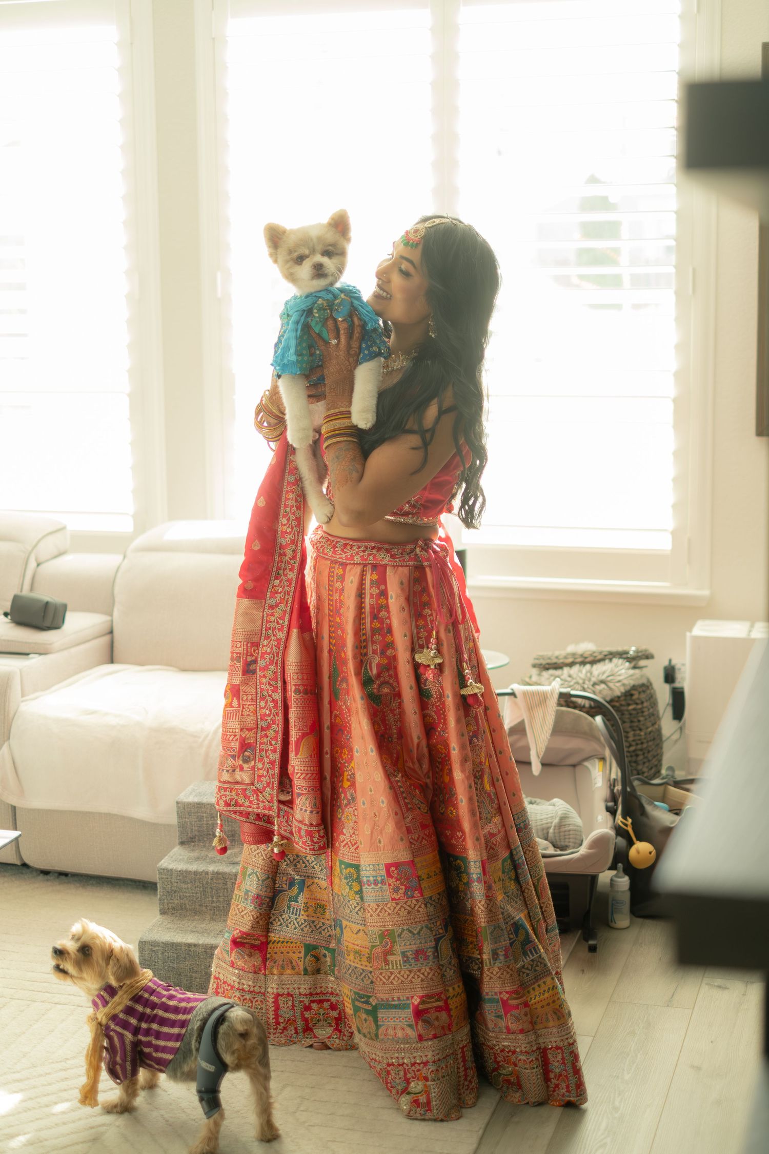 Bride in pink lehenga holds small dog while another dog stands nearby in bright indoor setting.