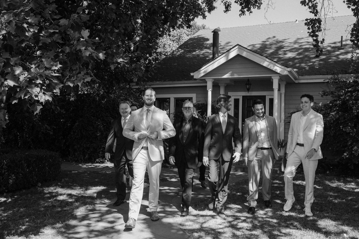 Black and white photo of a groom and groomsmen walking confidently outdoors near a white house.