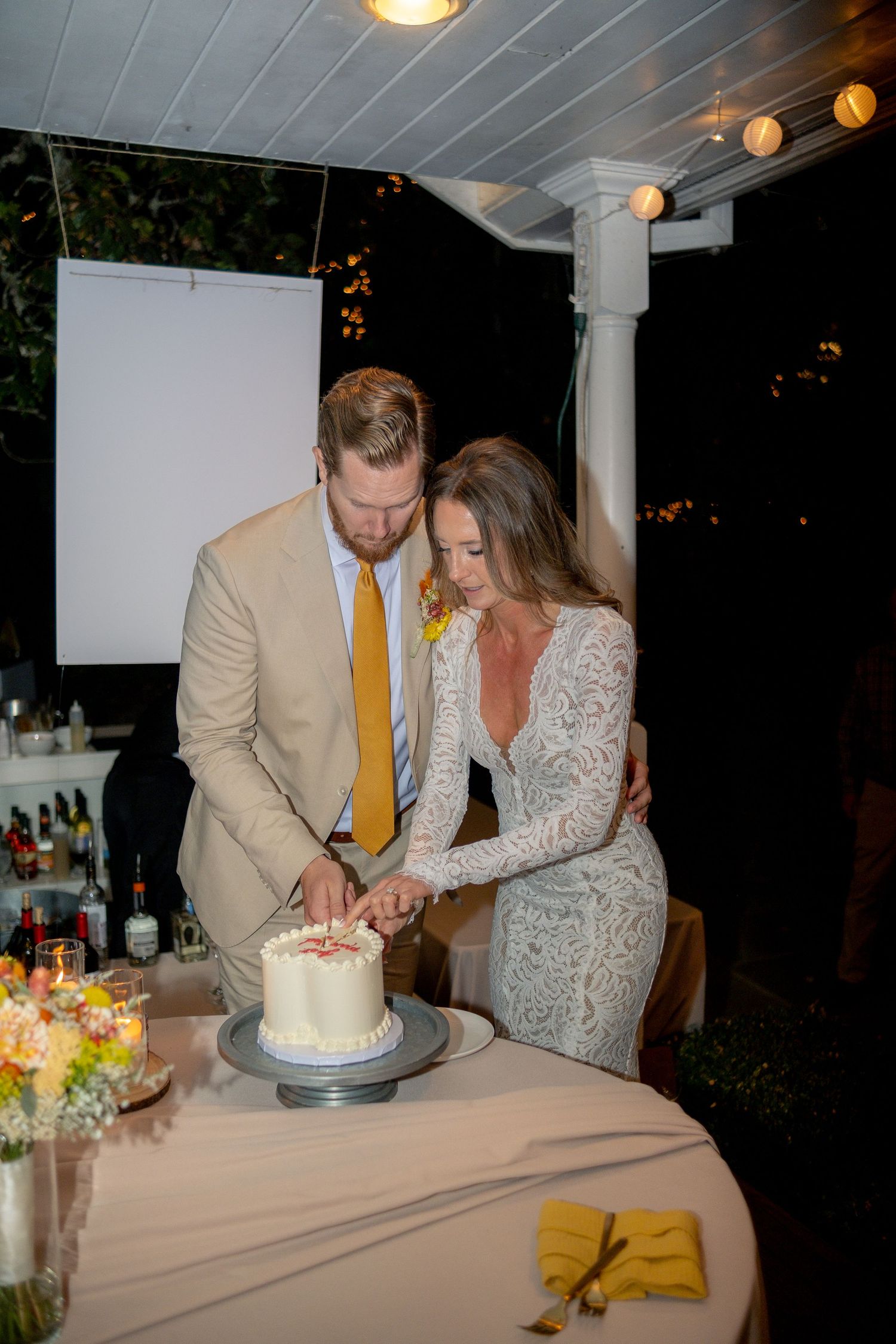 Bride and groom cutting white wedding cake together at outdoor evening reception with string lights.