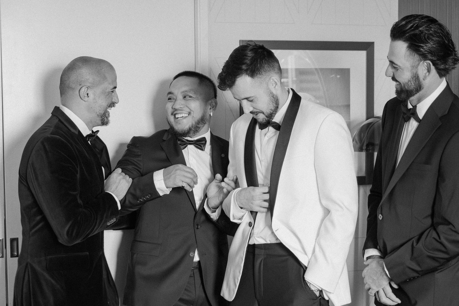 Groomsmen in tuxedos laughing and helping each other get dressed before a wedding ceremony.