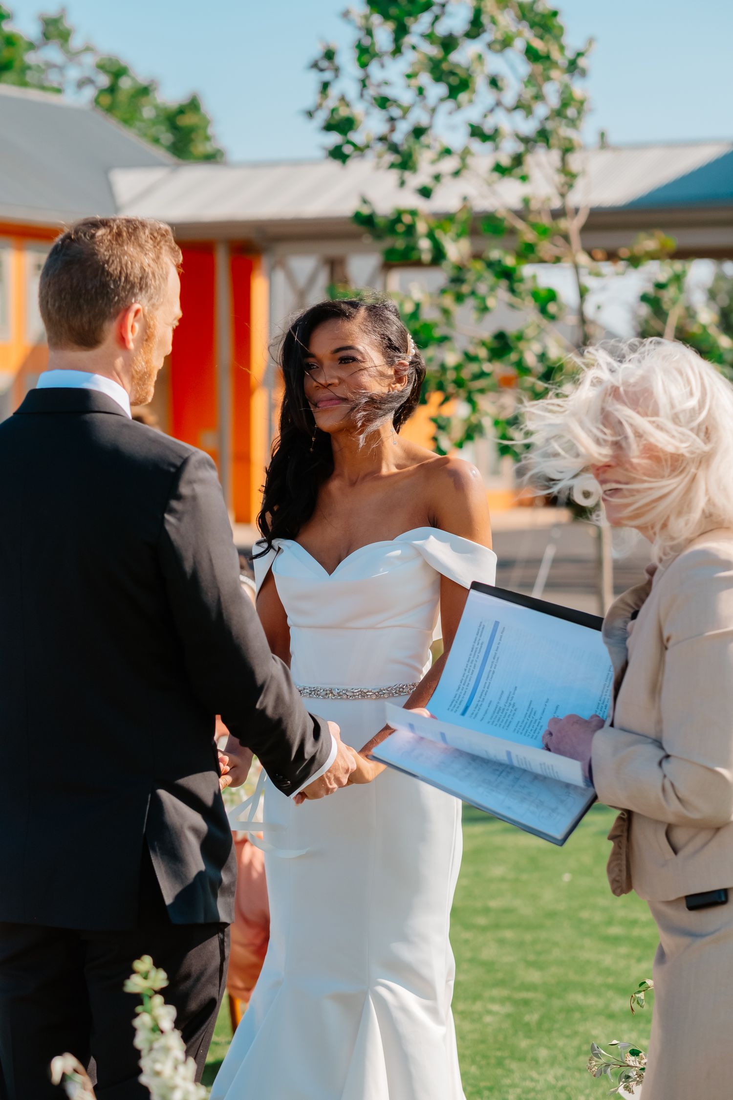 Bride in off-shoulder white gown smiles at groom during outdoor wedding ceremony with officiant.