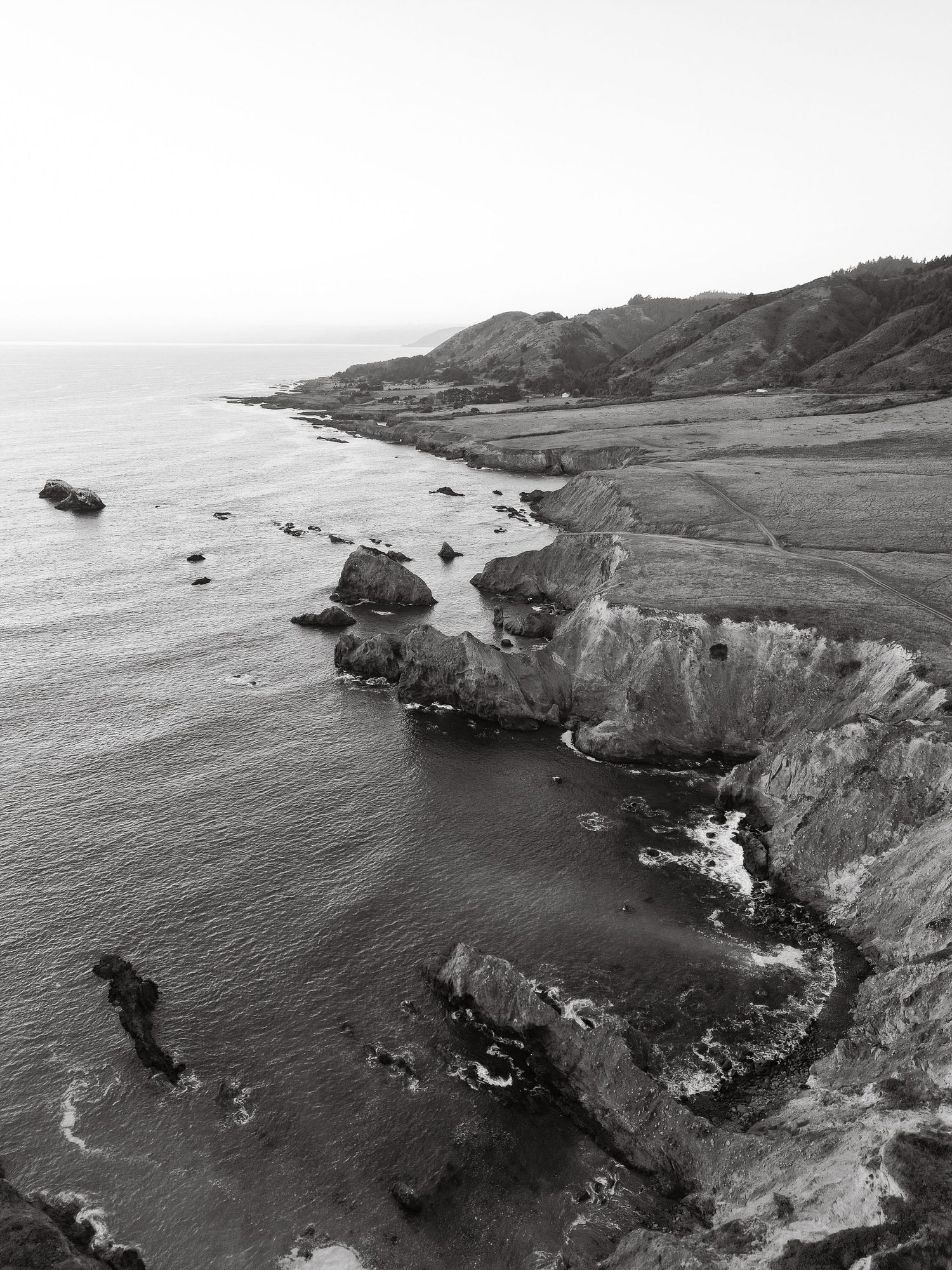 Black and white aerial view of a rugged rocky coastline with cliffs, ocean waves, and rolling hills.