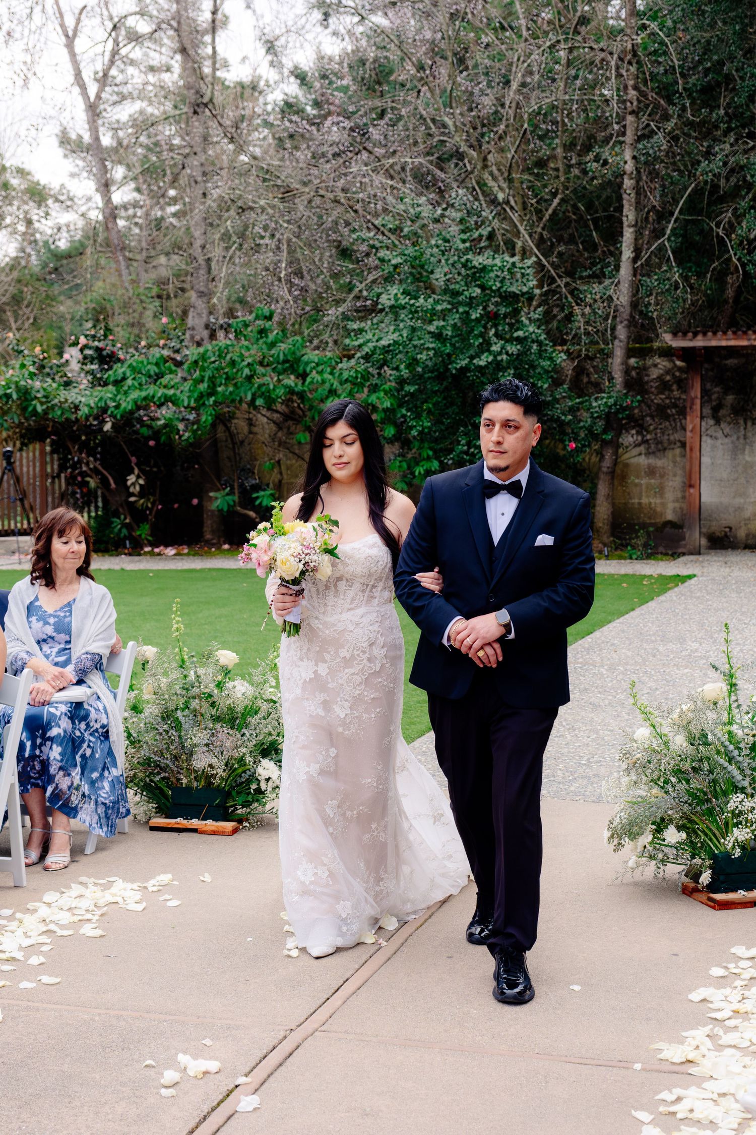 Bride in lace gown walks down outdoor aisle with escort at garden wedding ceremony.