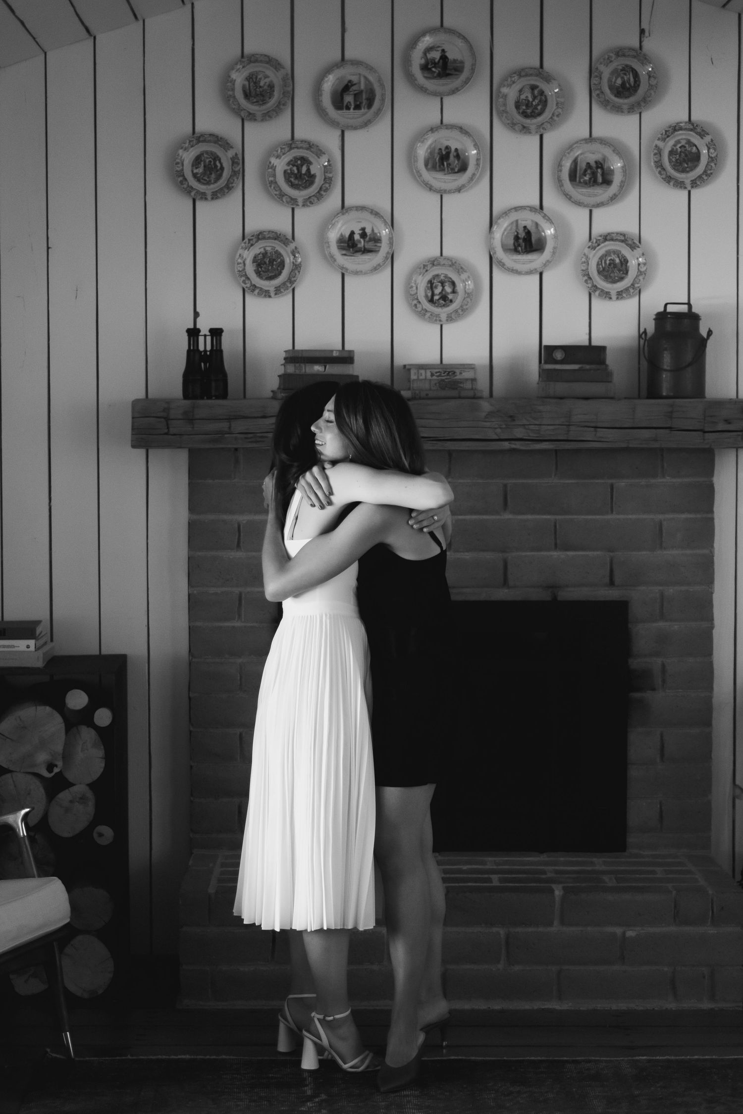 Two women embracing in a hug by a brick fireplace with decorative plates on the wall in a black and white photo.