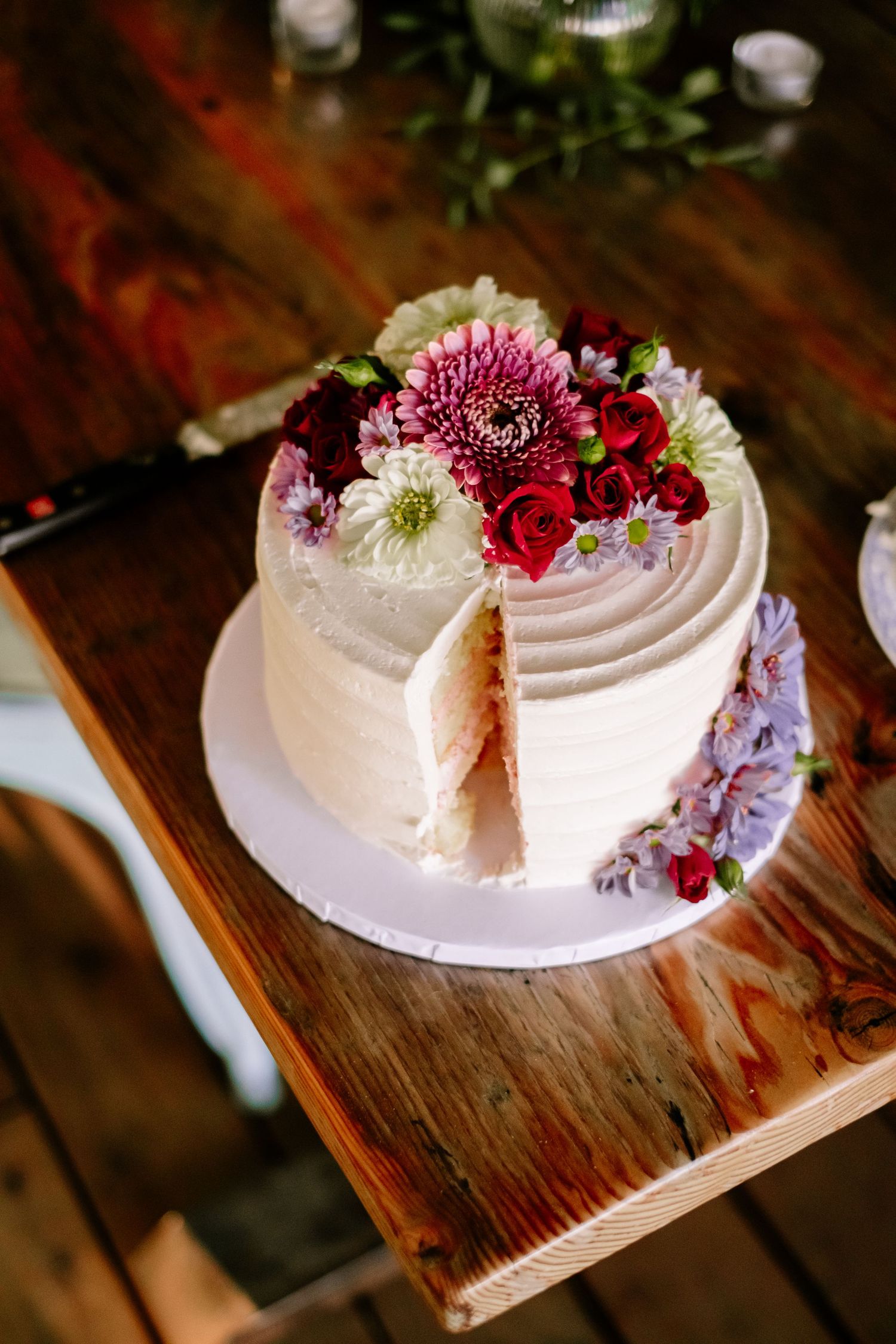White frosted single-tier wedding cake topped with fresh pink, red, and white flowers on a rustic wooden table.