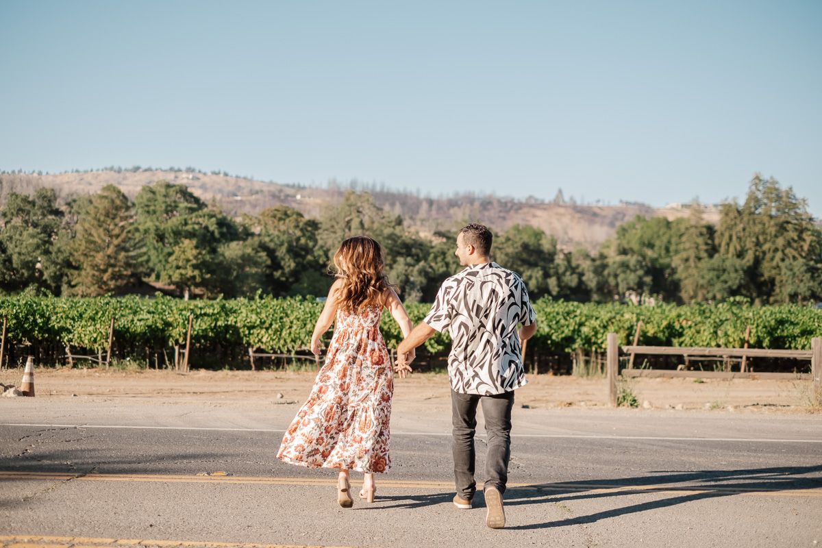 Couple holding hands walking through a sunny vineyard landscape with rolling hills in background.