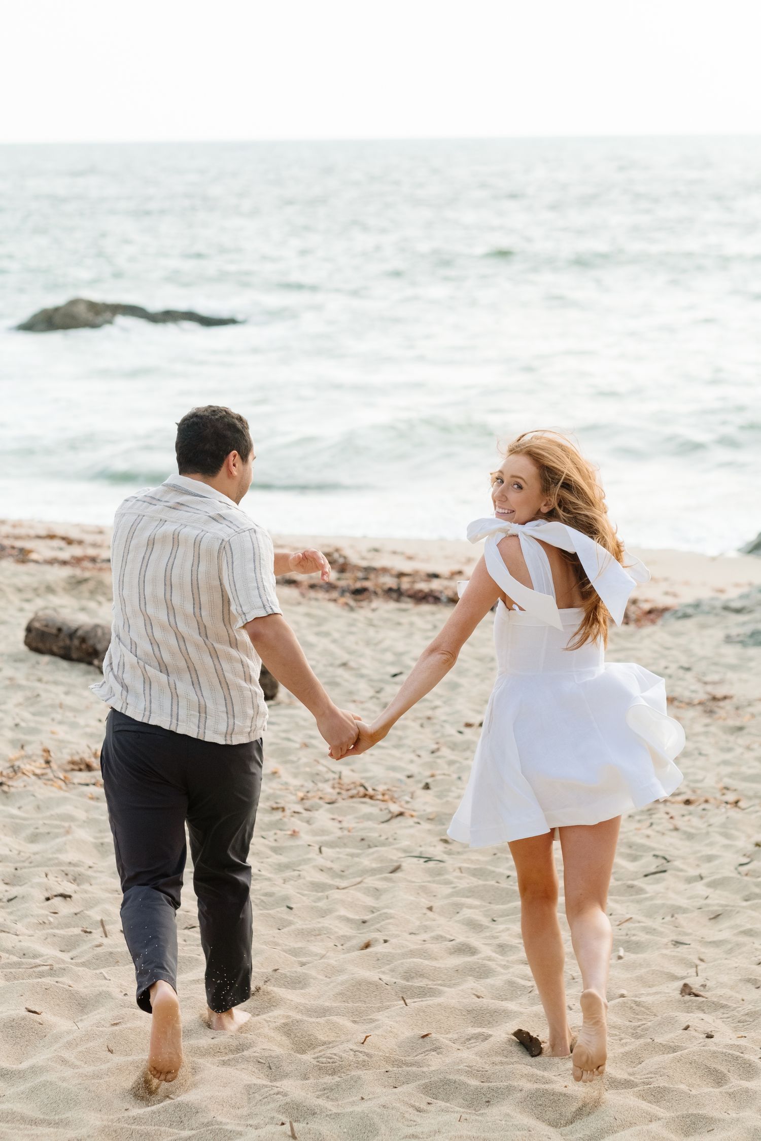 Couple holding hands walking barefoot on sandy beach toward ocean waves, woman in white dress looking back over shoulder.