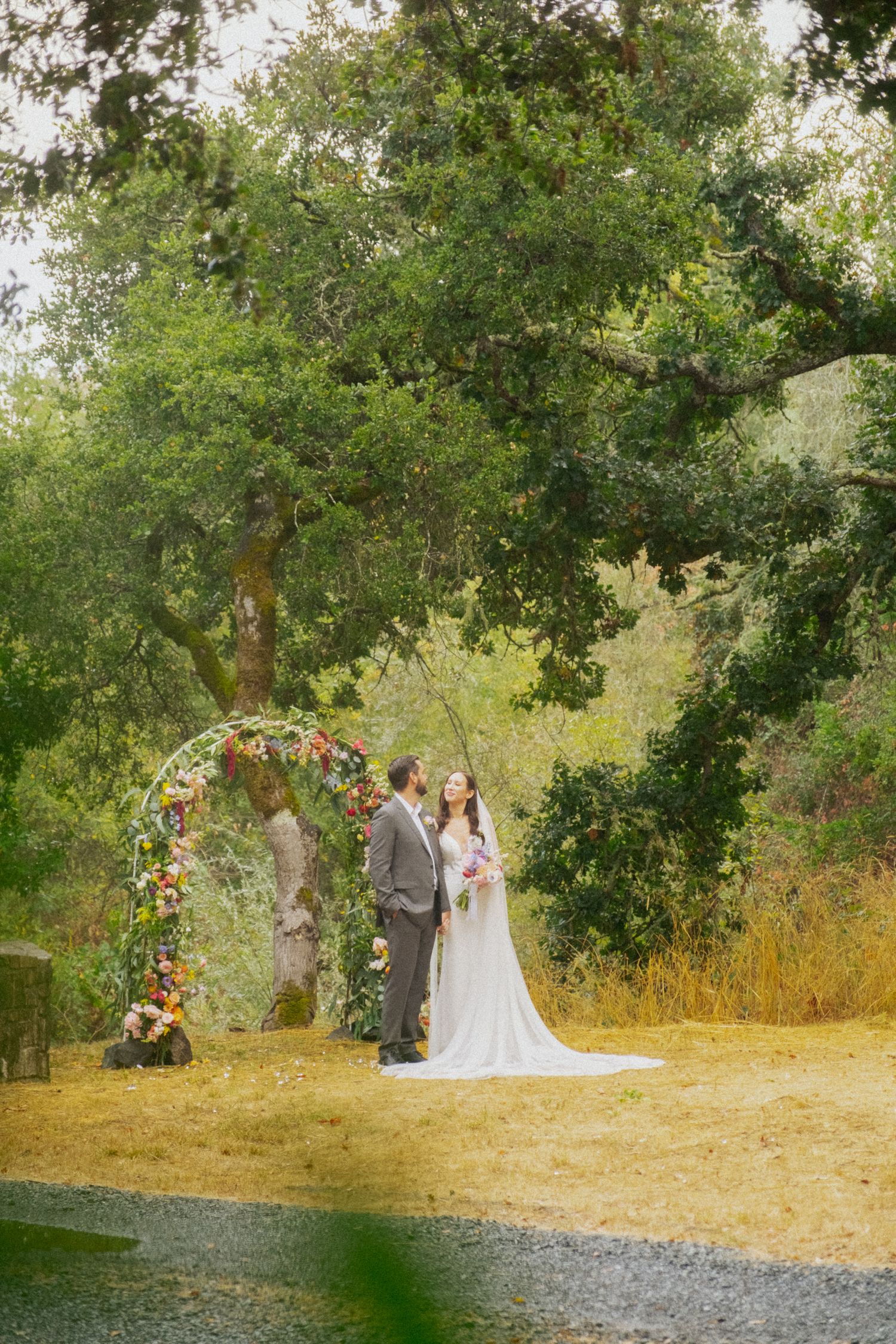 Bride and groom exchange vows under a colorful floral arch surrounded by lush green trees outdoors.