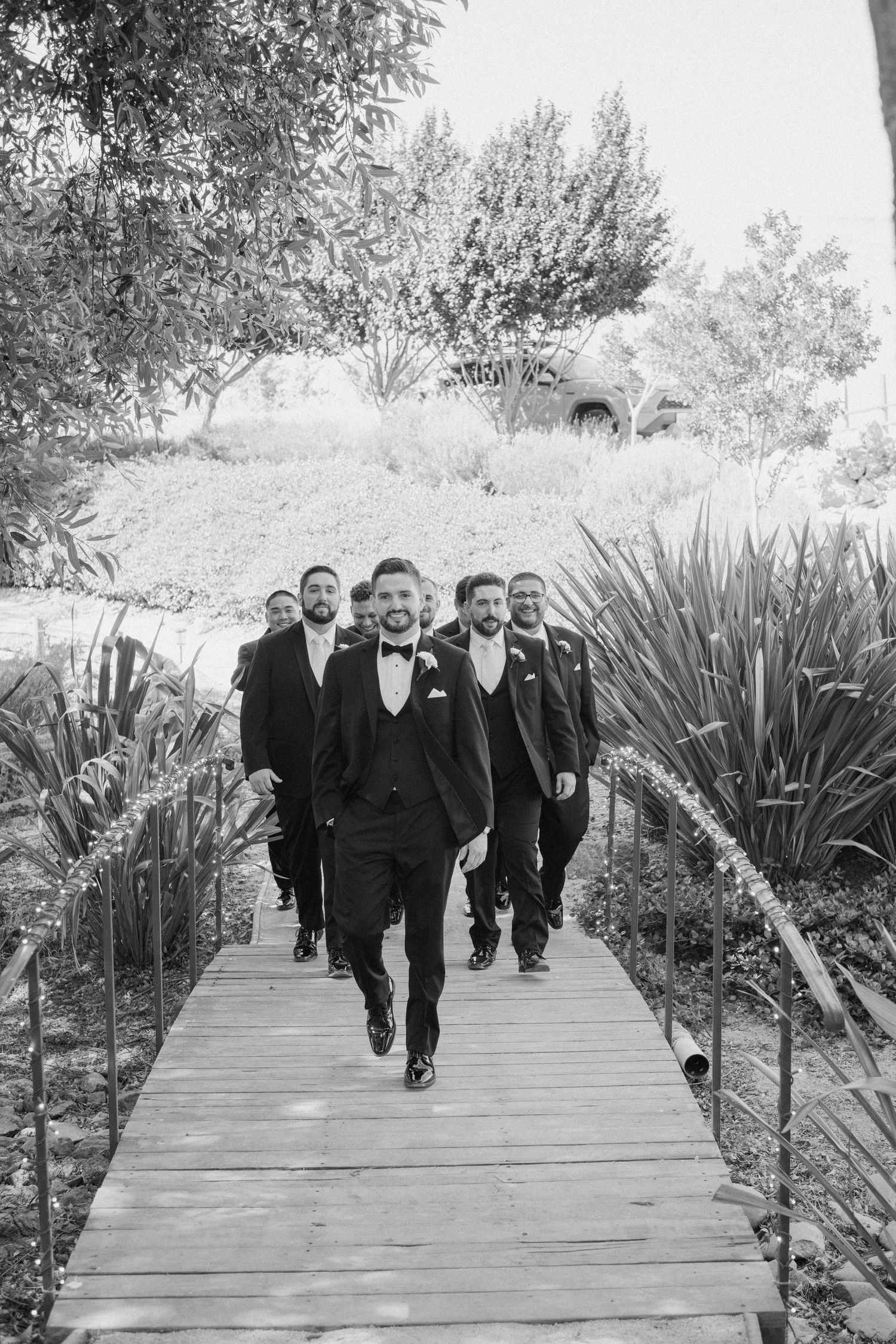 Black and white photo of a dapper groom leading his groomsmen across a wooden bridge outdoors.