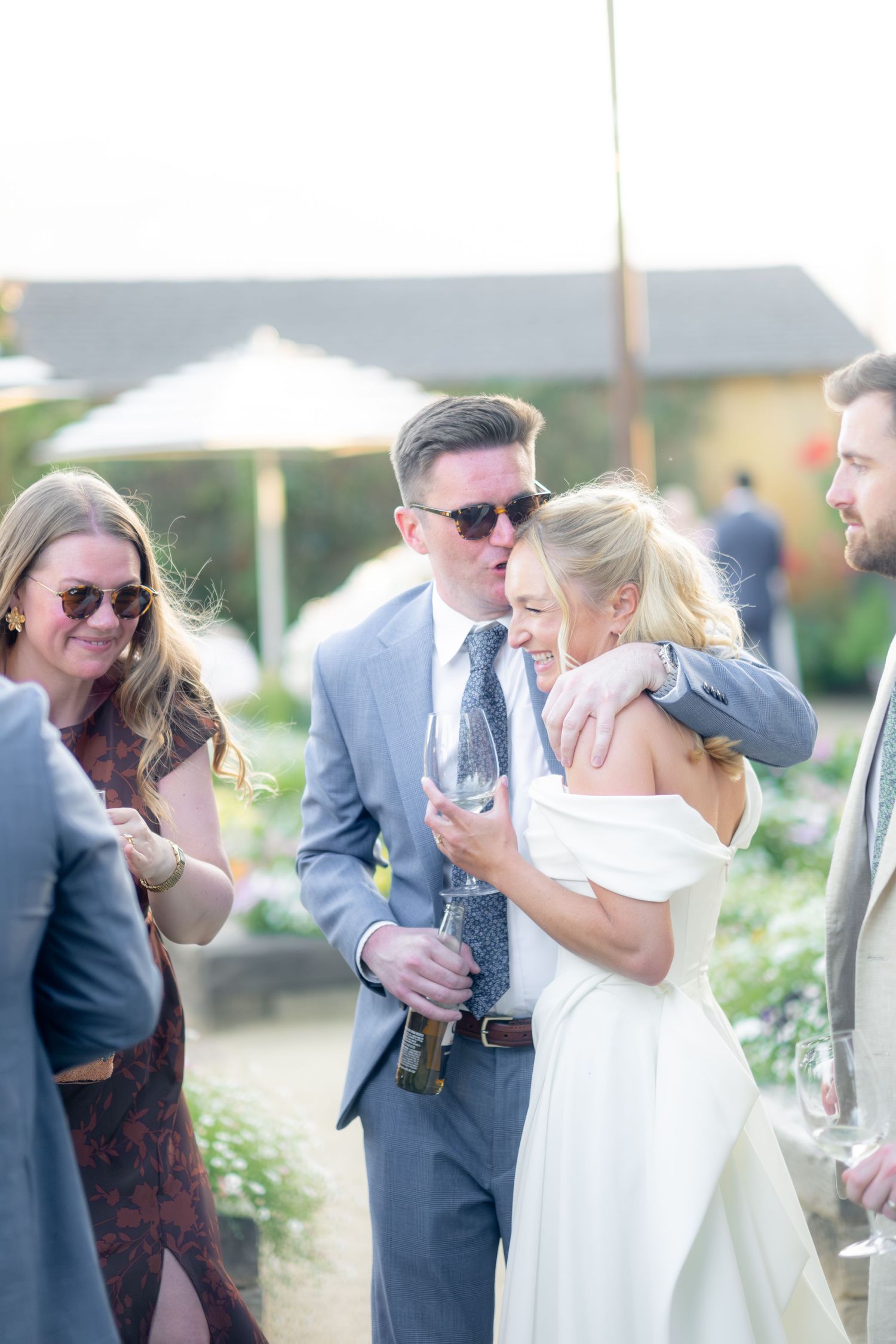 Bride in off-shoulder white gown embracing groom in grey suit with sunglasses during outdoor wedding cocktail hour.