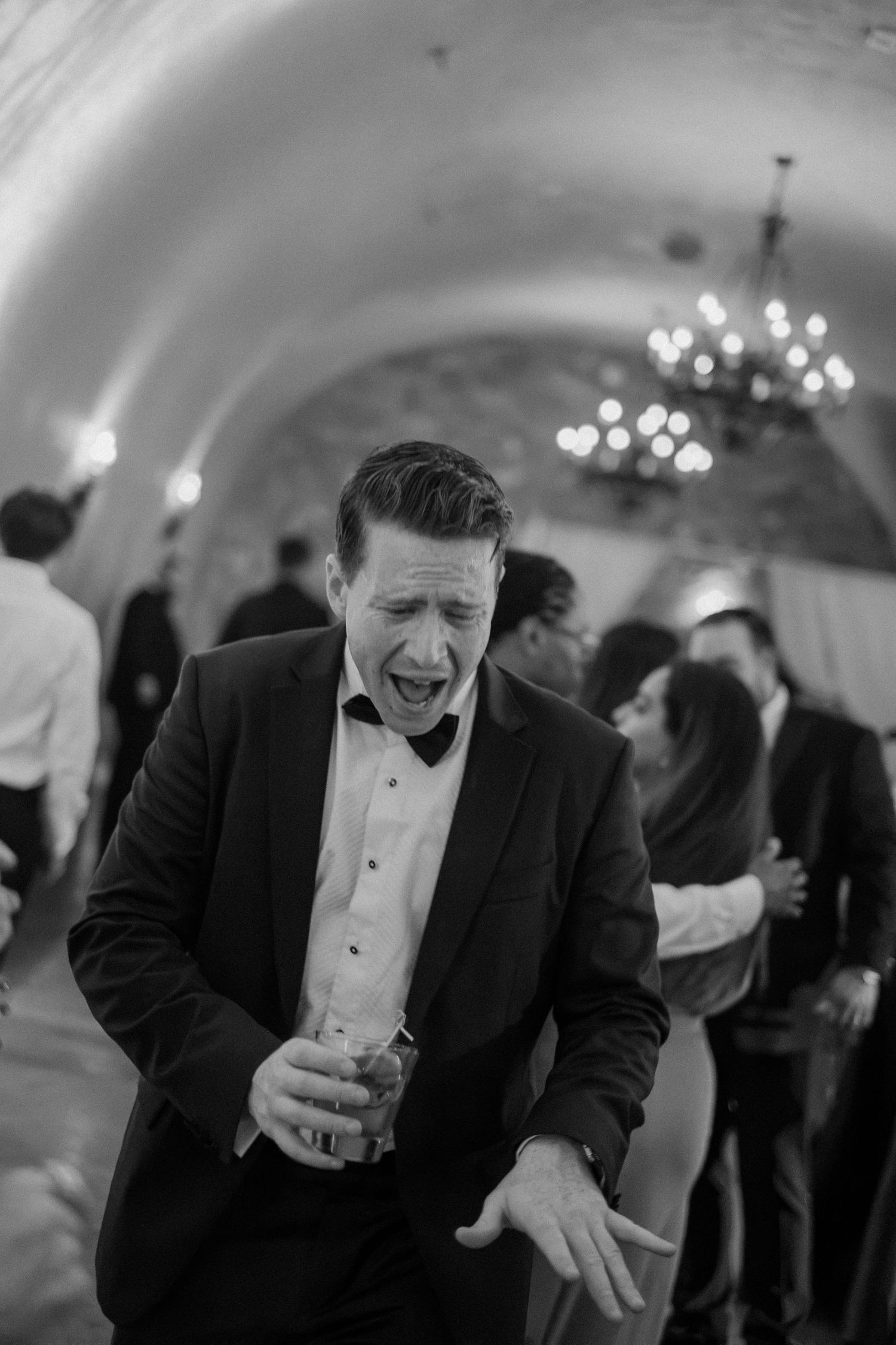 Black and white photo of wedding guest dancing energetically with drink in hand at reception.