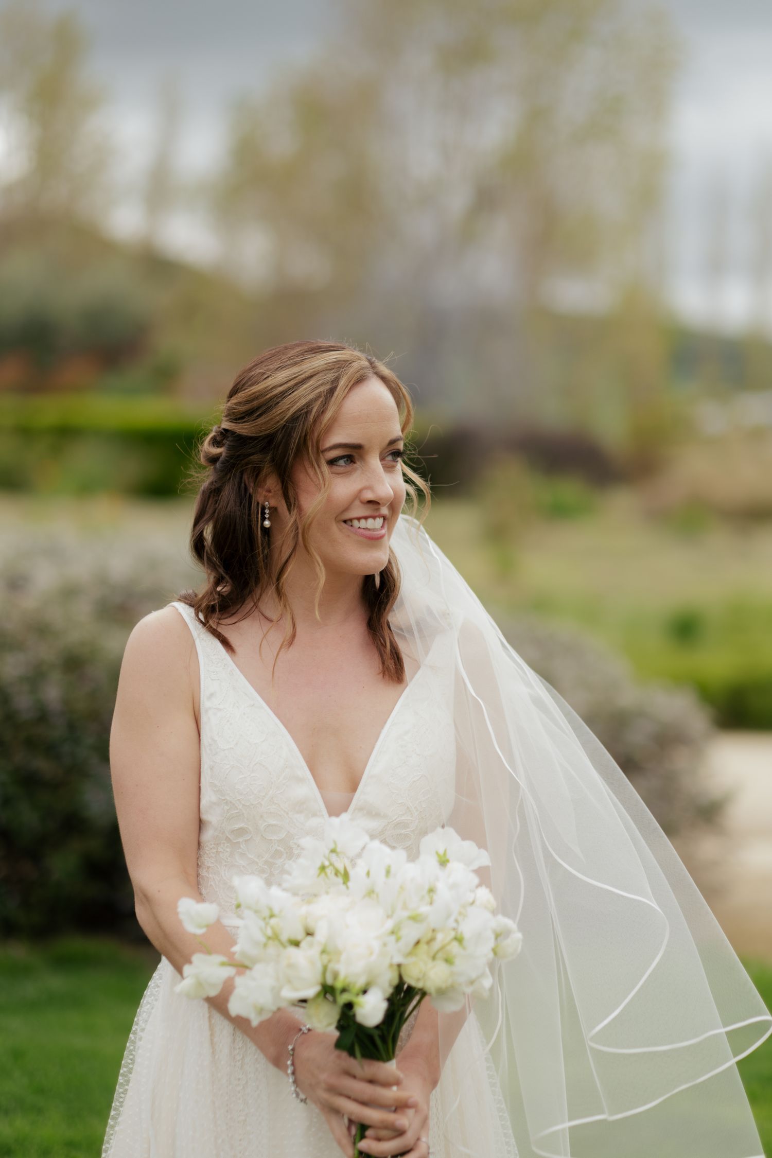 Radiant bride in deep V-neck lace wedding gown holding white bouquet with veil flowing in the breeze.